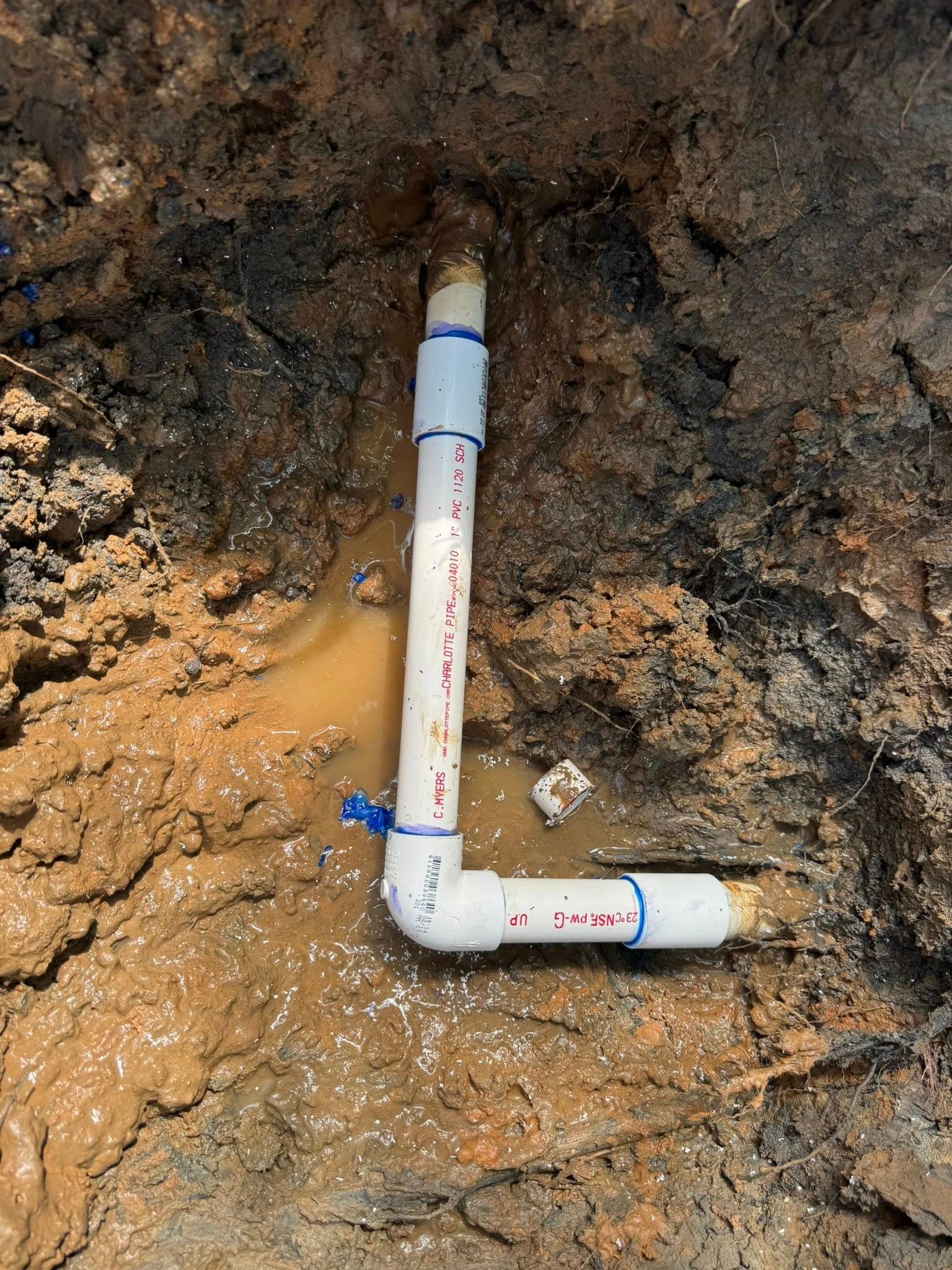 A white PVC pipe assembly with a 90-degree elbow connector installed in a muddy, excavated trench in the ground.