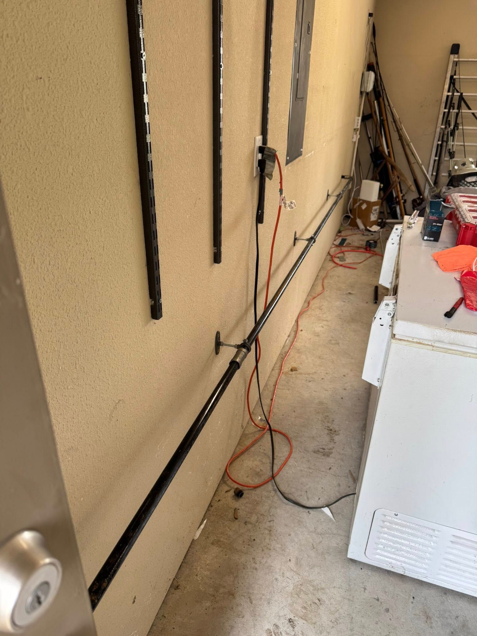 A garage wall with a horizontal black power strip mounted, vertical wall tracks, and a red extension cord on the floor.