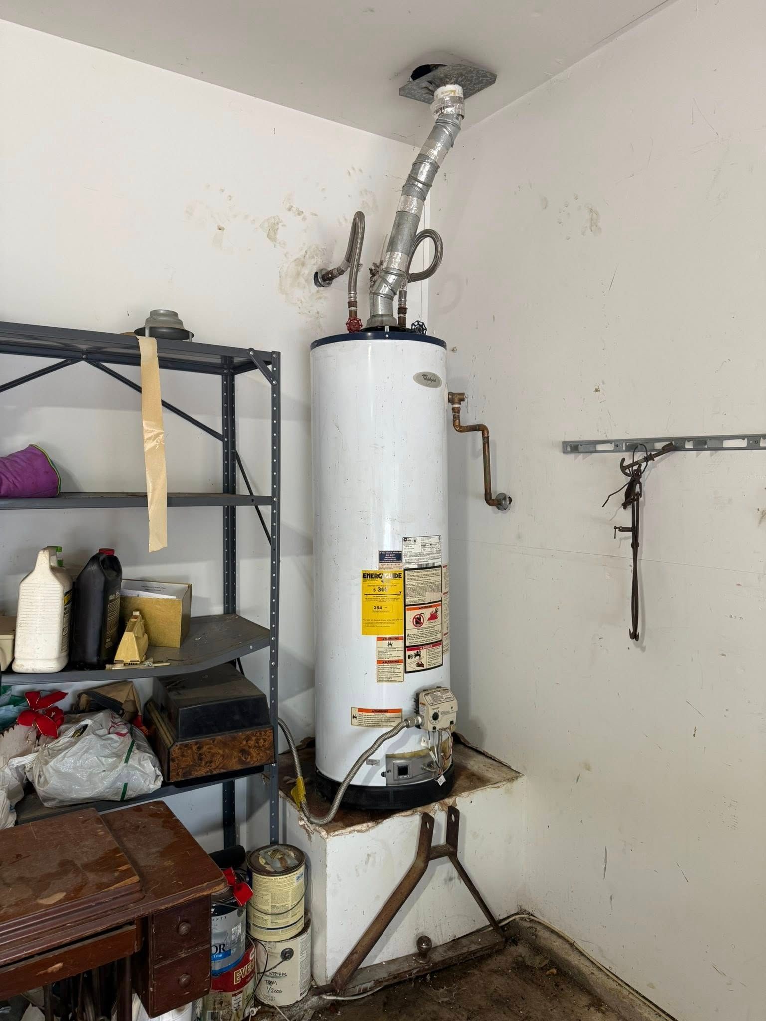 A white water heater stands in a corner on a metal platform, with a ventilation pipe extending up to the ceiling.