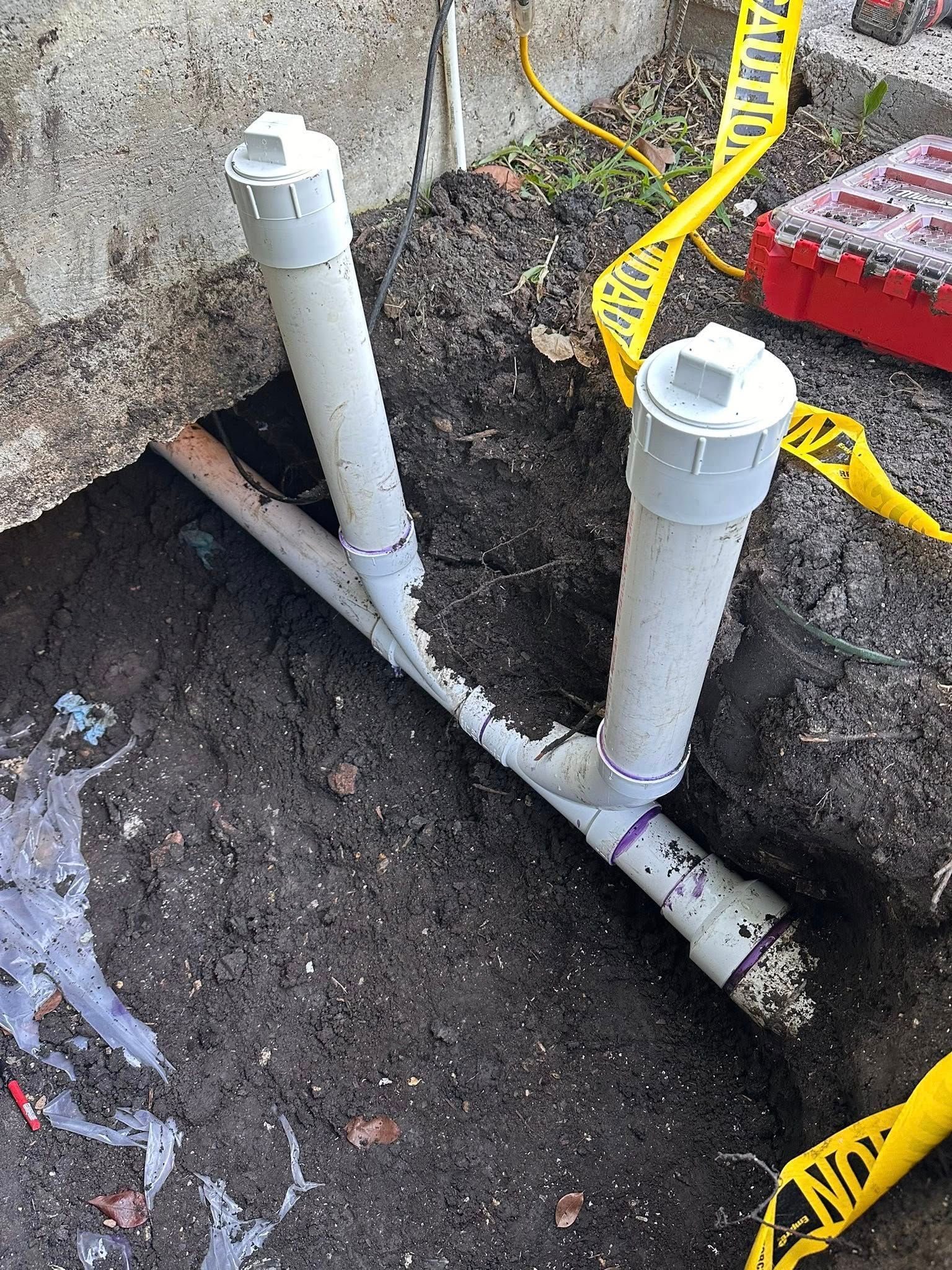 Two vertical PVC pipes with capped ends installed on a main drain line in an excavated dirt trench next to a concrete wall.