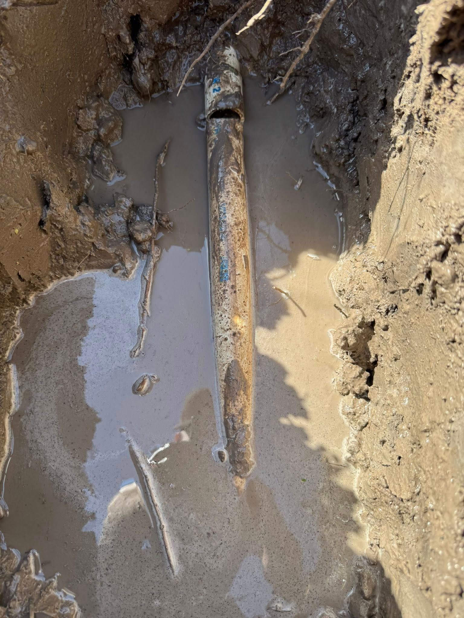 A rusted, cylindrical pipe partially submerged in muddy water within a narrow, dirt-walled trench.