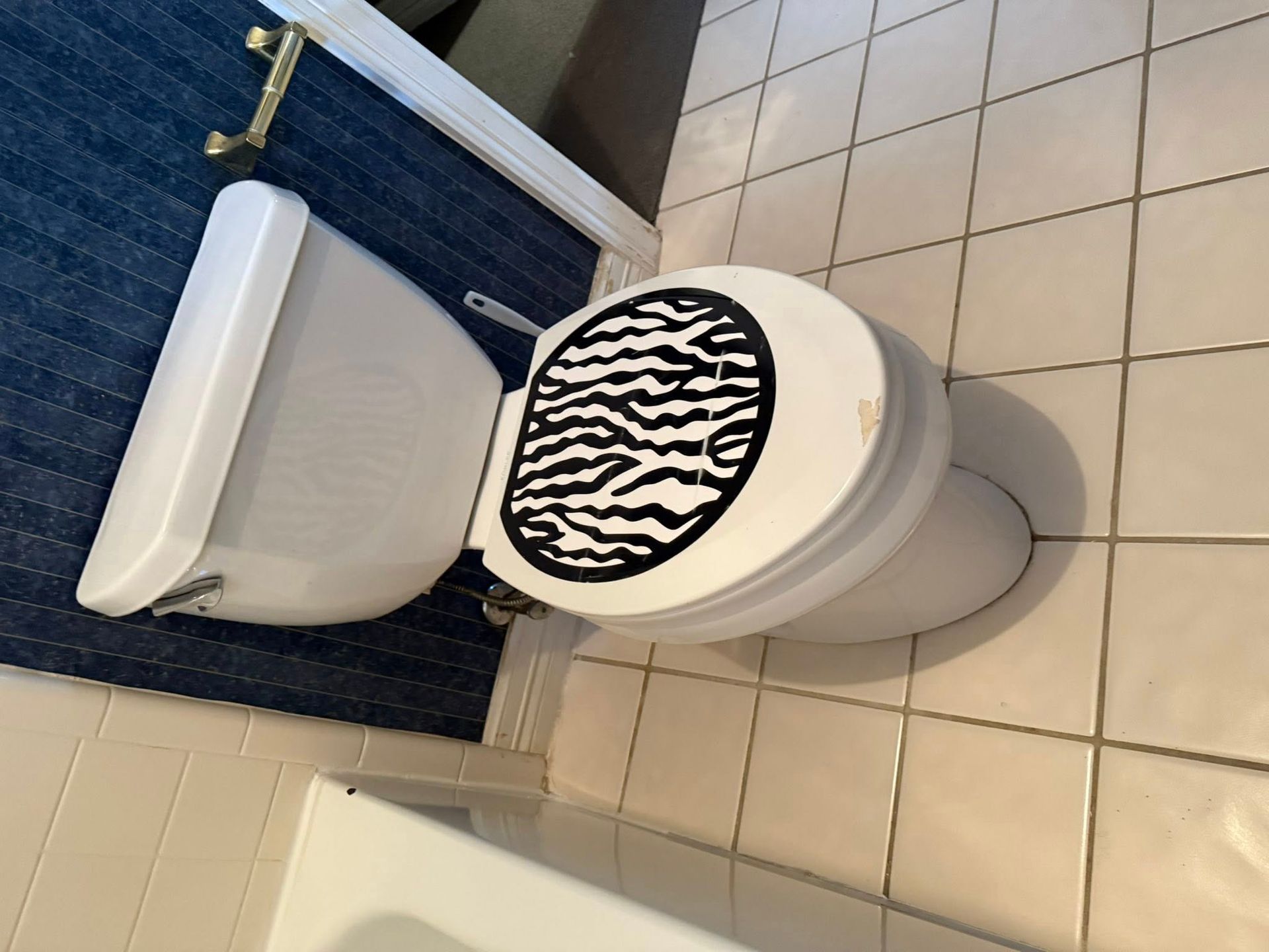 A white toilet with a black and white zebra-patterned toilet seat cover in a bathroom with blue wallpaper and tiled floors.