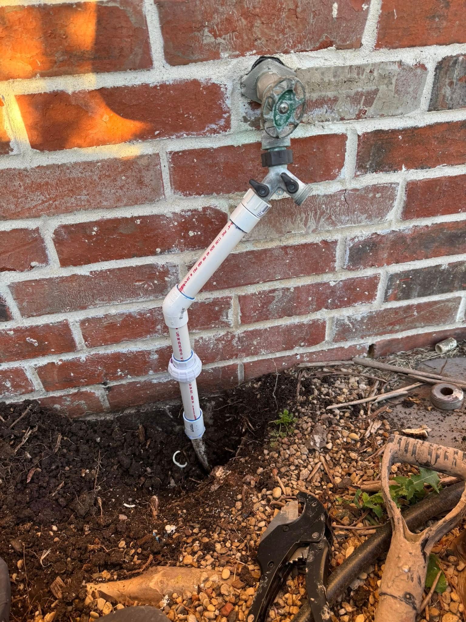 A spigot attached to a brick wall with an L-shaped PVC pipe extension extending into the soil below.