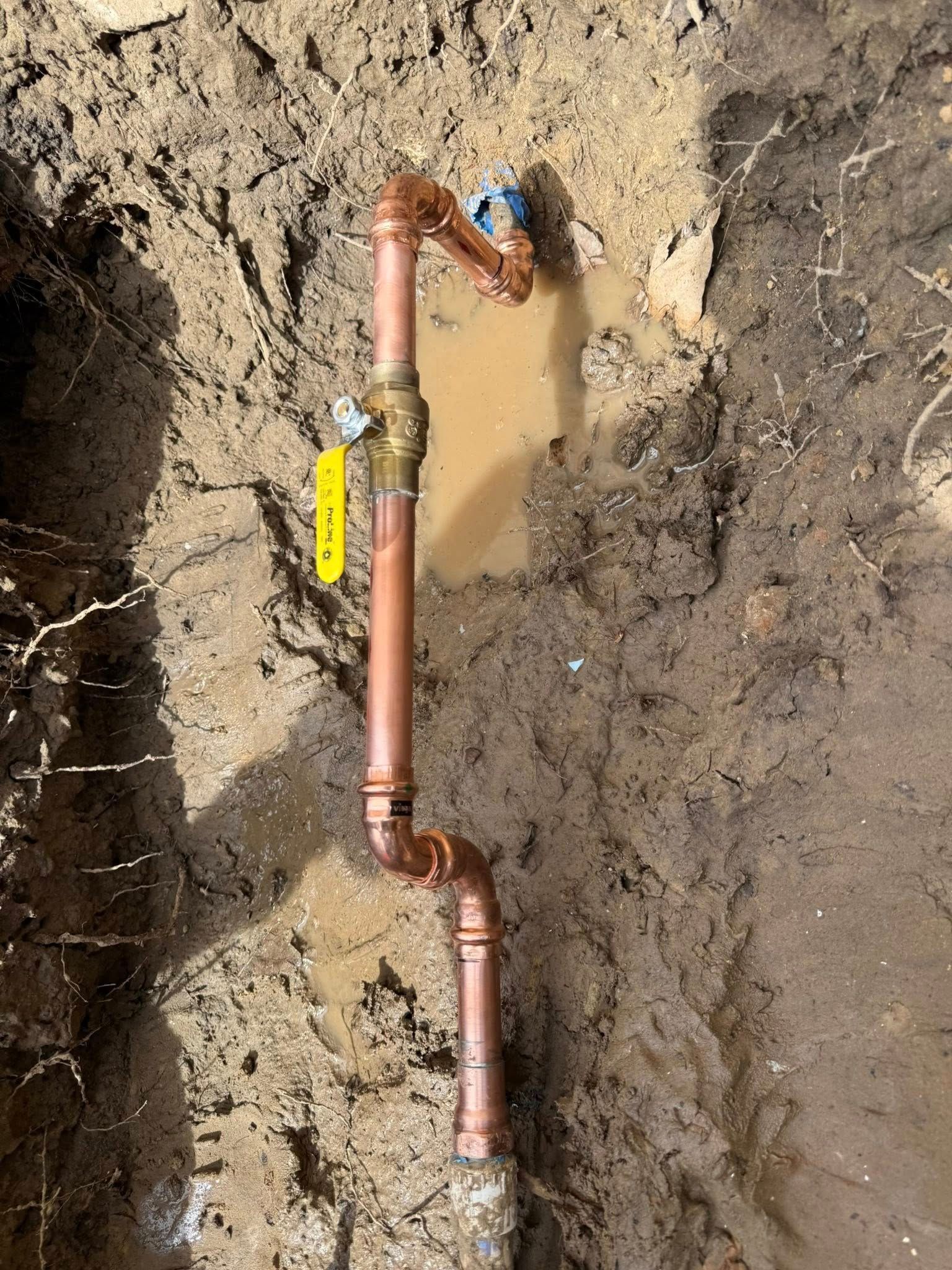A copper pipe assembly with a ball valve and yellow handle installed in muddy, excavated ground.