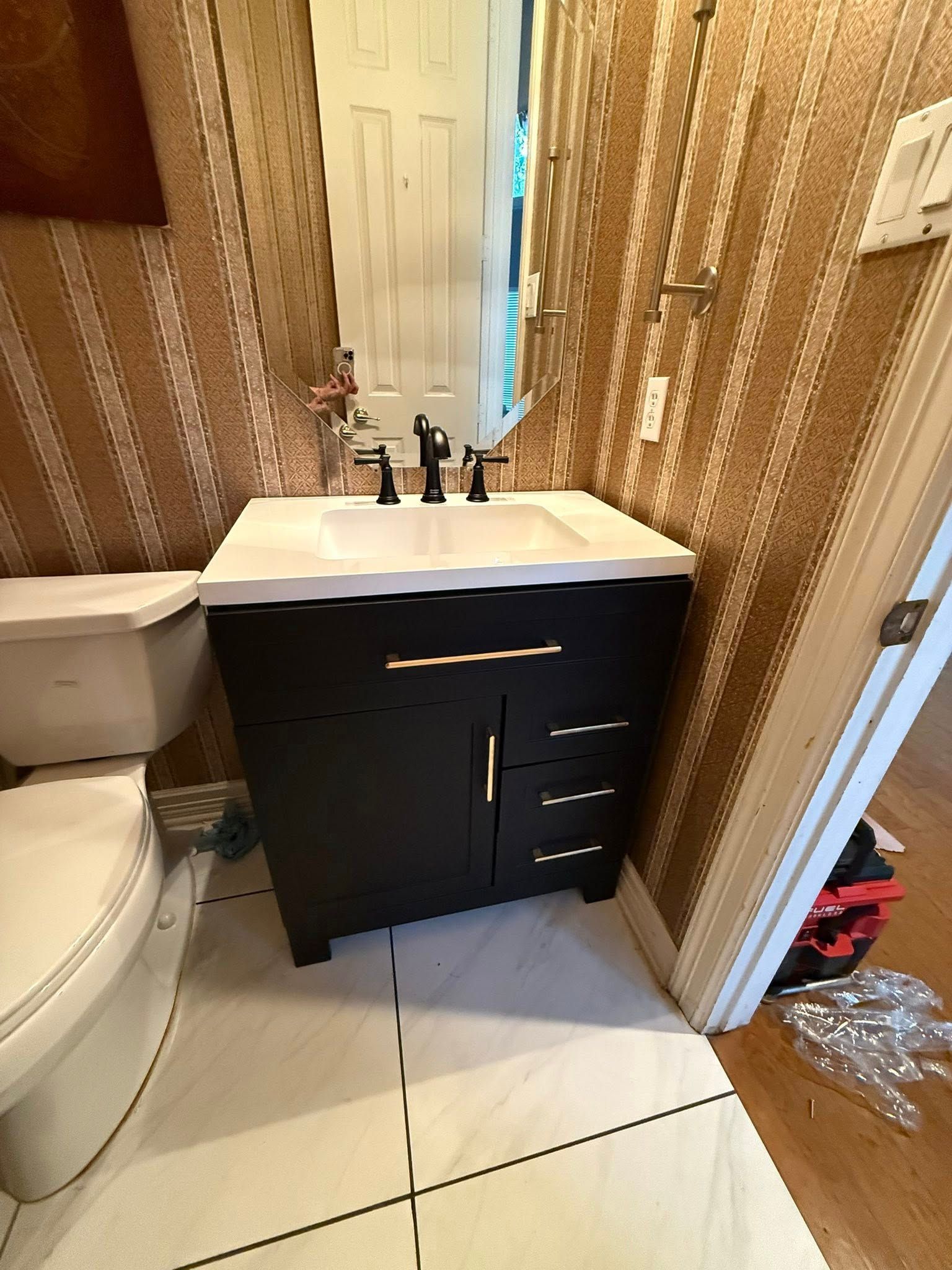 A black vanity with a white countertop and faucet sits between a toilet and a doorway in a room with patterned wallpaper.