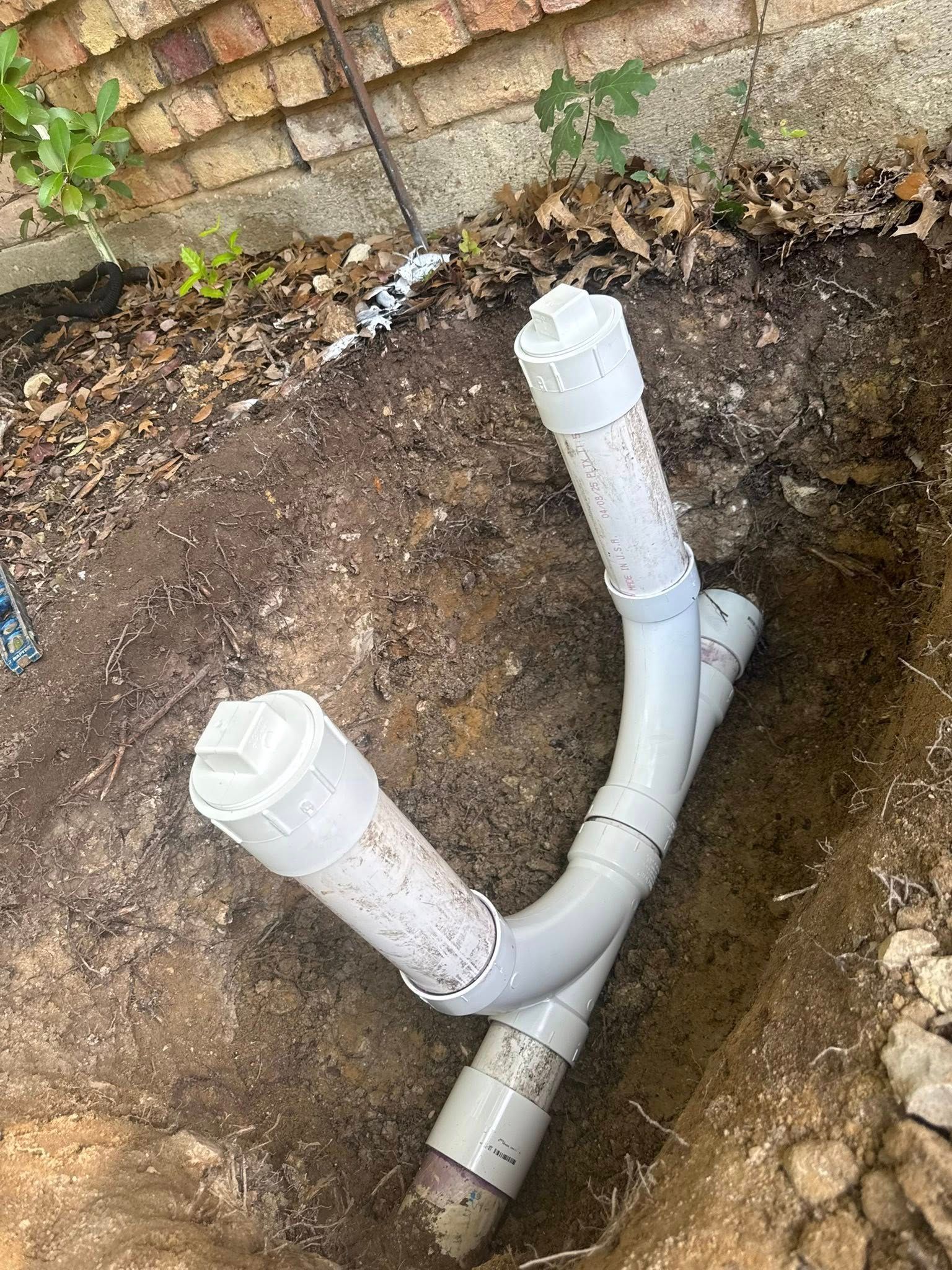Two white PVC cleanout pipes connected by a Y-fitting, partially buried in a dirt trench next to a brick wall.