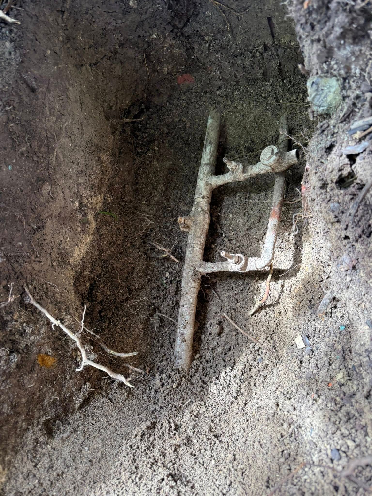 A rusted metal frame with two parallel vertical rods and crossbars partially buried in a narrow dirt trench.