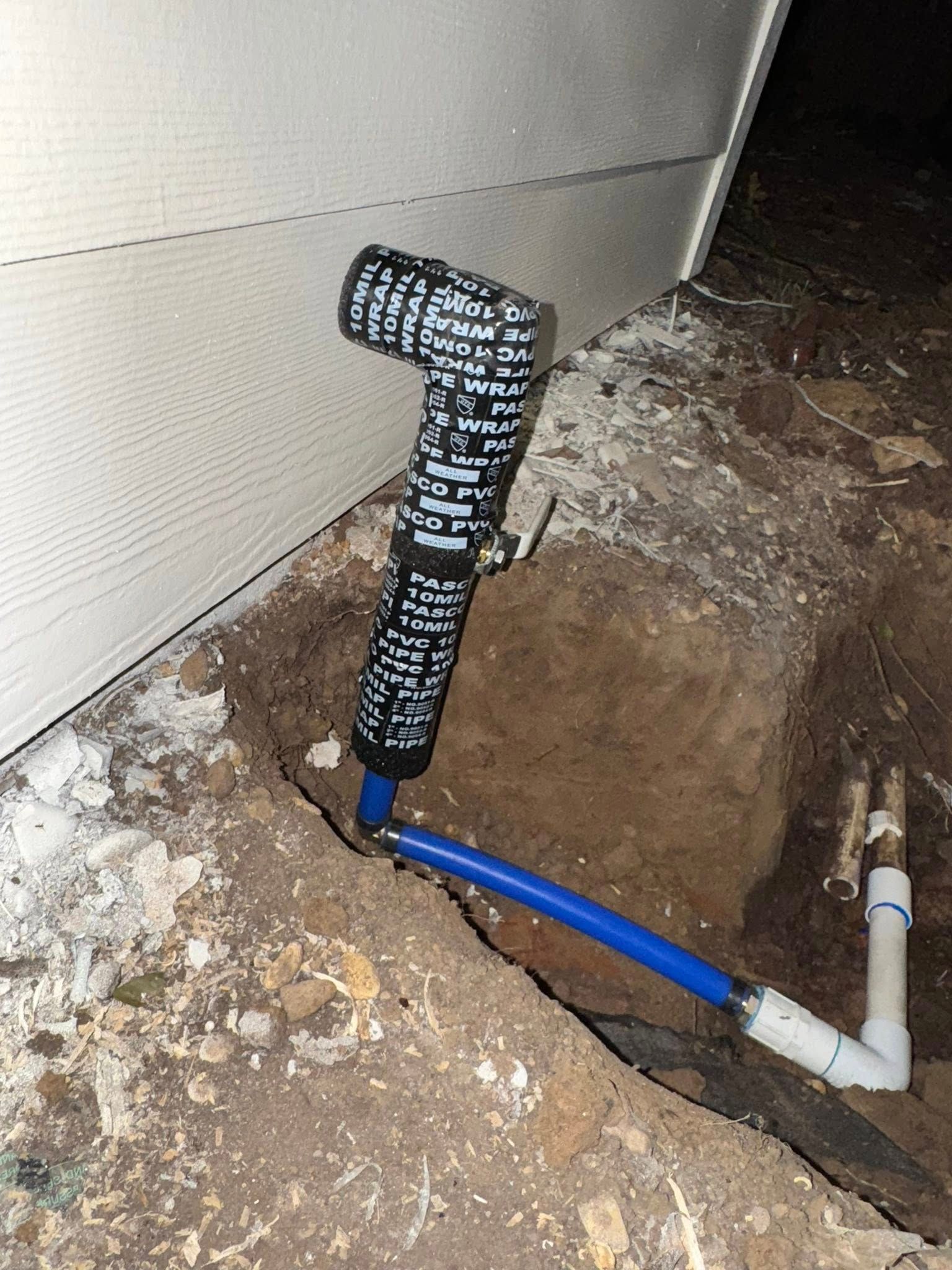 An outdoor water pipe riser with a valve, wrapped in black foam insulation, connected to blue and white PVC piping.