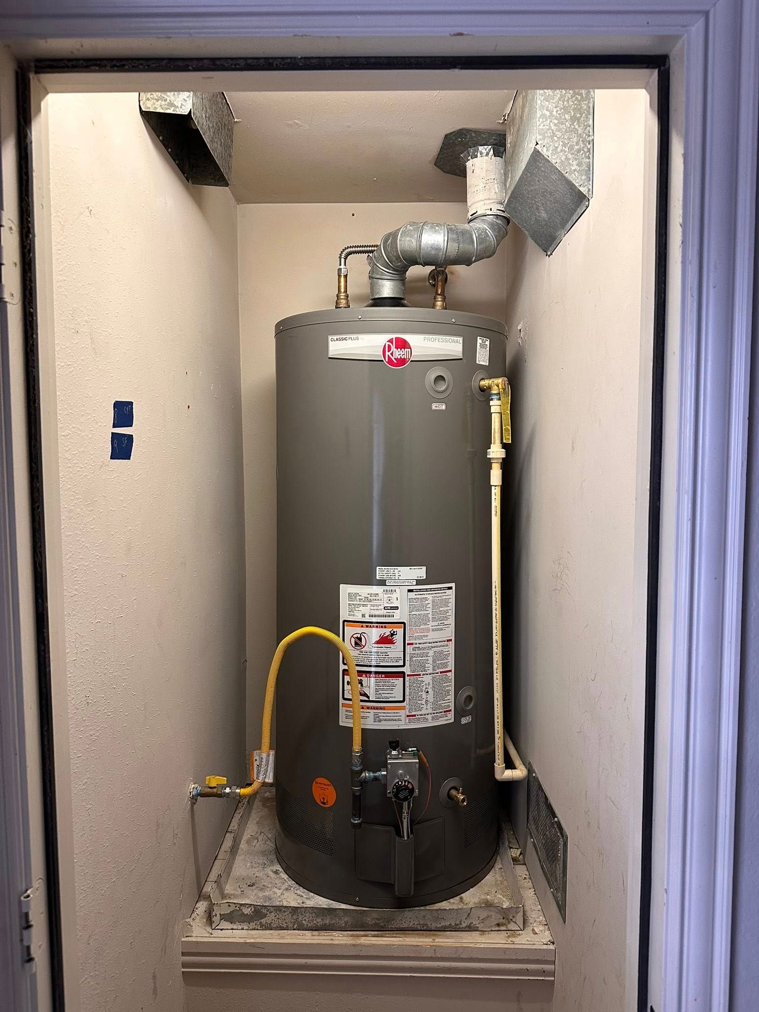 A grey water heater sits in a closet on a drainage pan, connected to gas and water lines with an exhaust vent above.