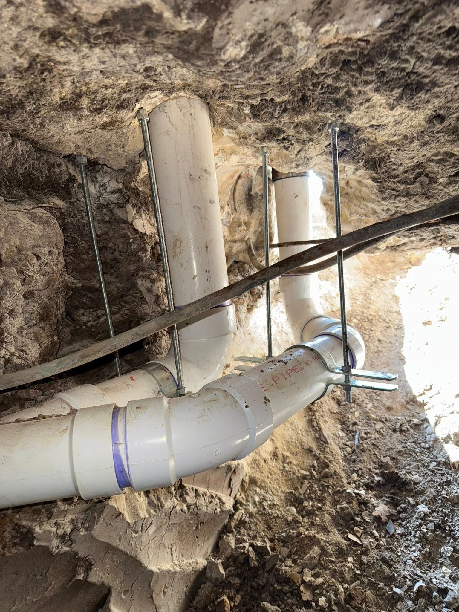 White PVC plumbing pipes, joined by fittings, are suspended by threaded rods in an excavated dirt trench.