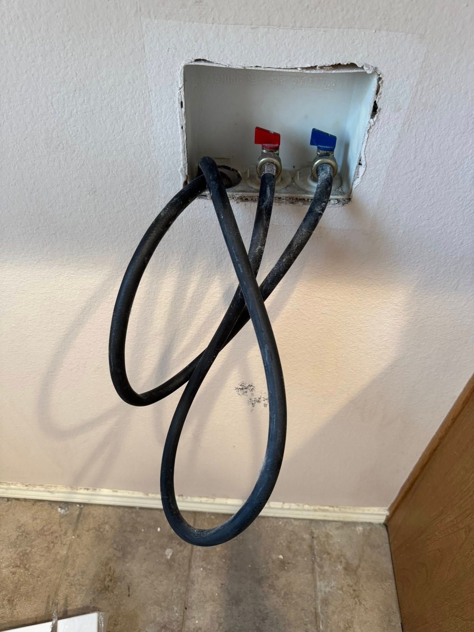 A laundry wall box with hot and cold water supply valves connected to two black rubber hoses hanging downward.