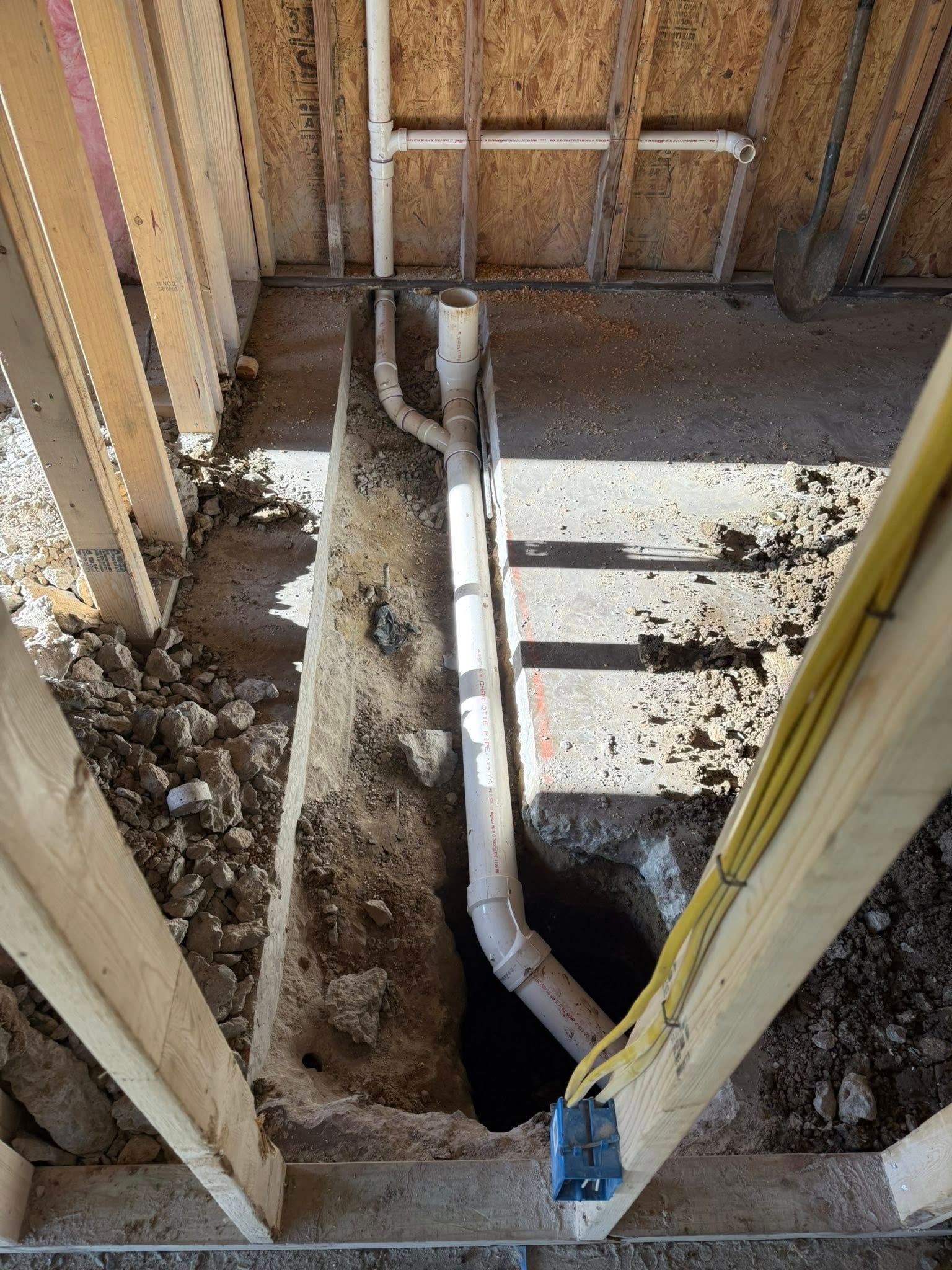 An unfinished room featuring exposed wall studs and a white PVC drain pipe running through a trench in a concrete floor.
