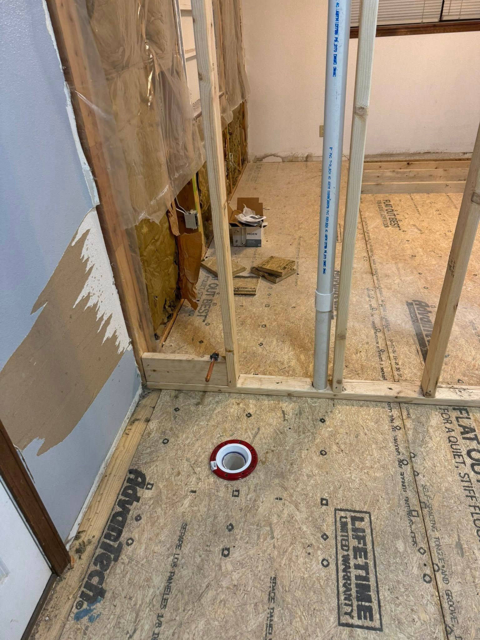 An interior view of a room under construction, showing wooden wall studs, insulation, a vertical pipe, and a floor drain.