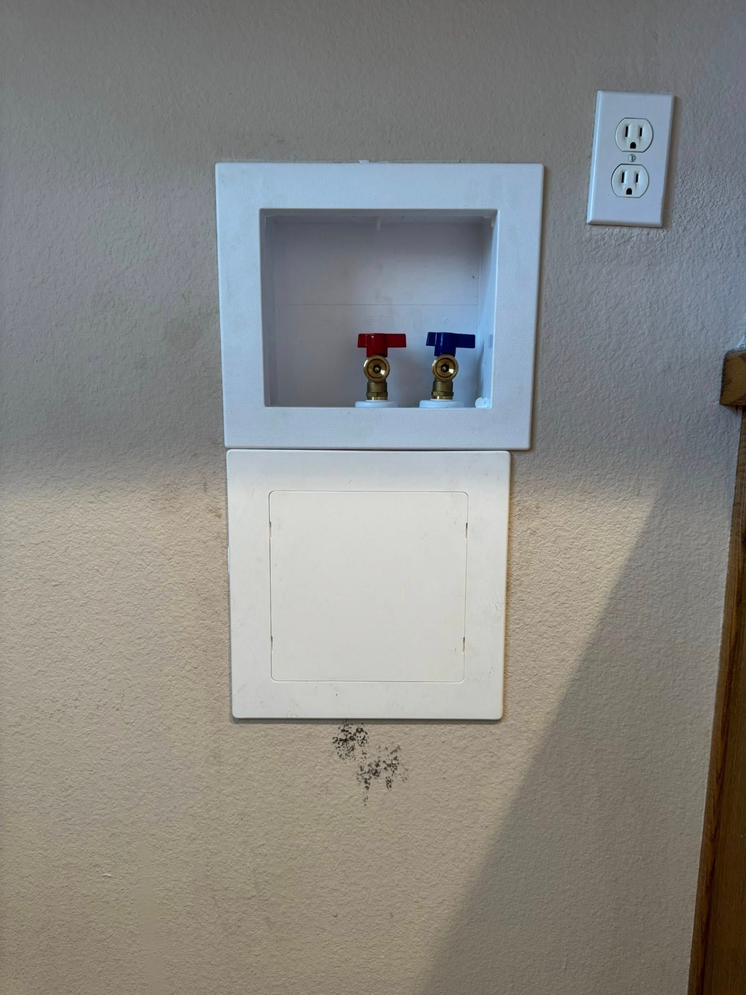 A laundry room wall box with hot and cold water valves, featuring a white cover plate below and an electrical outlet.