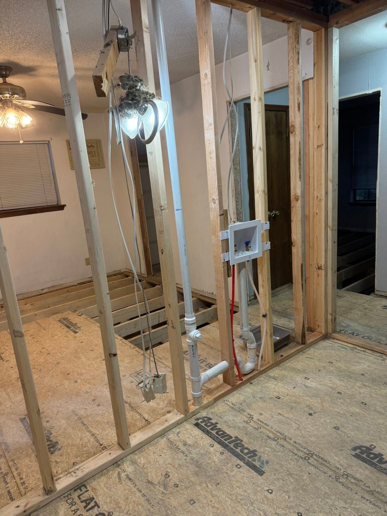 Construction site featuring exposed wooden wall studs, installed plumbing pipes, and a laundry box utility outlet.