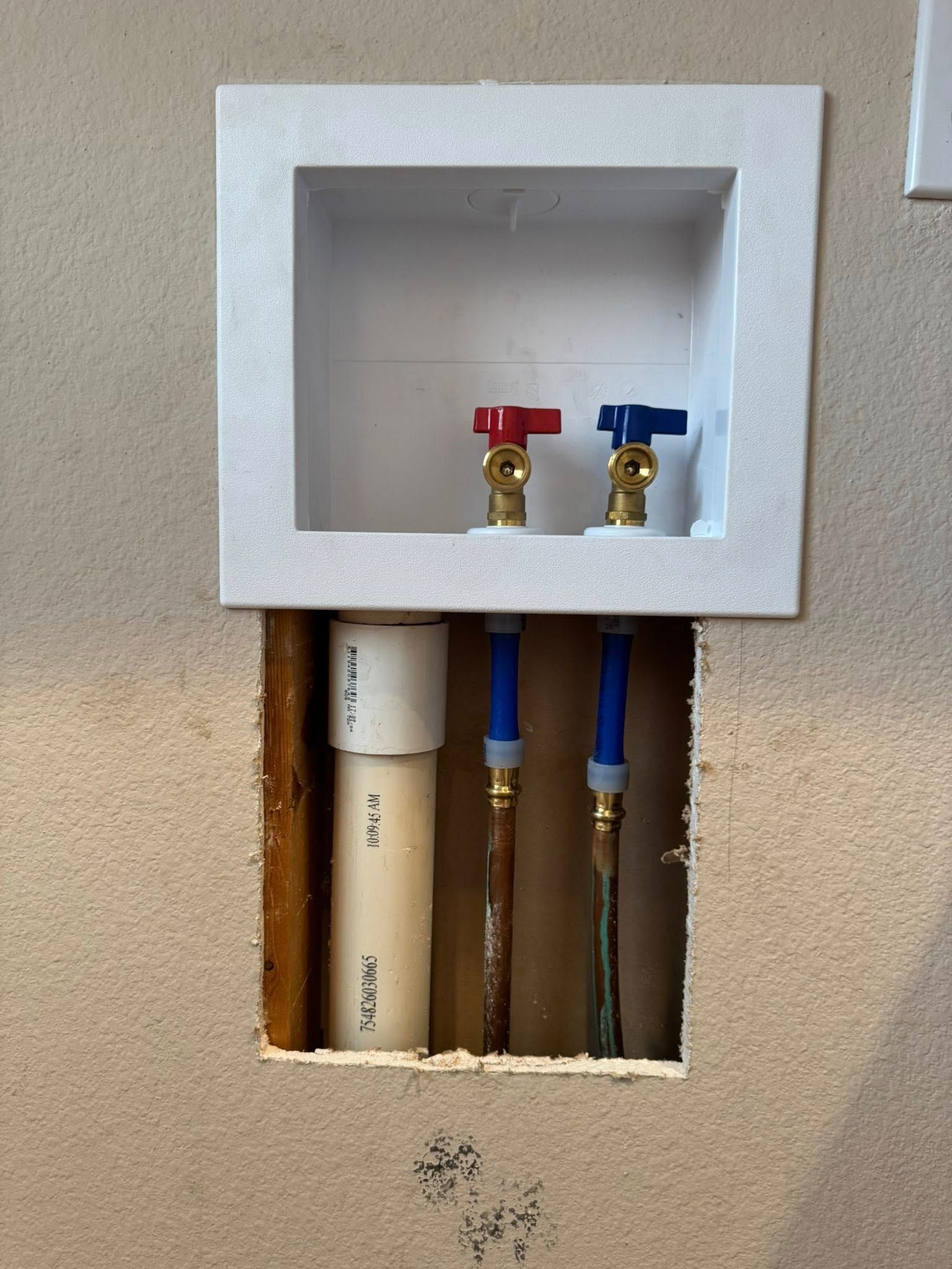 A white recessed washing machine utility box with red and blue water valves and a PVC drain pipe in an open wall cutout.