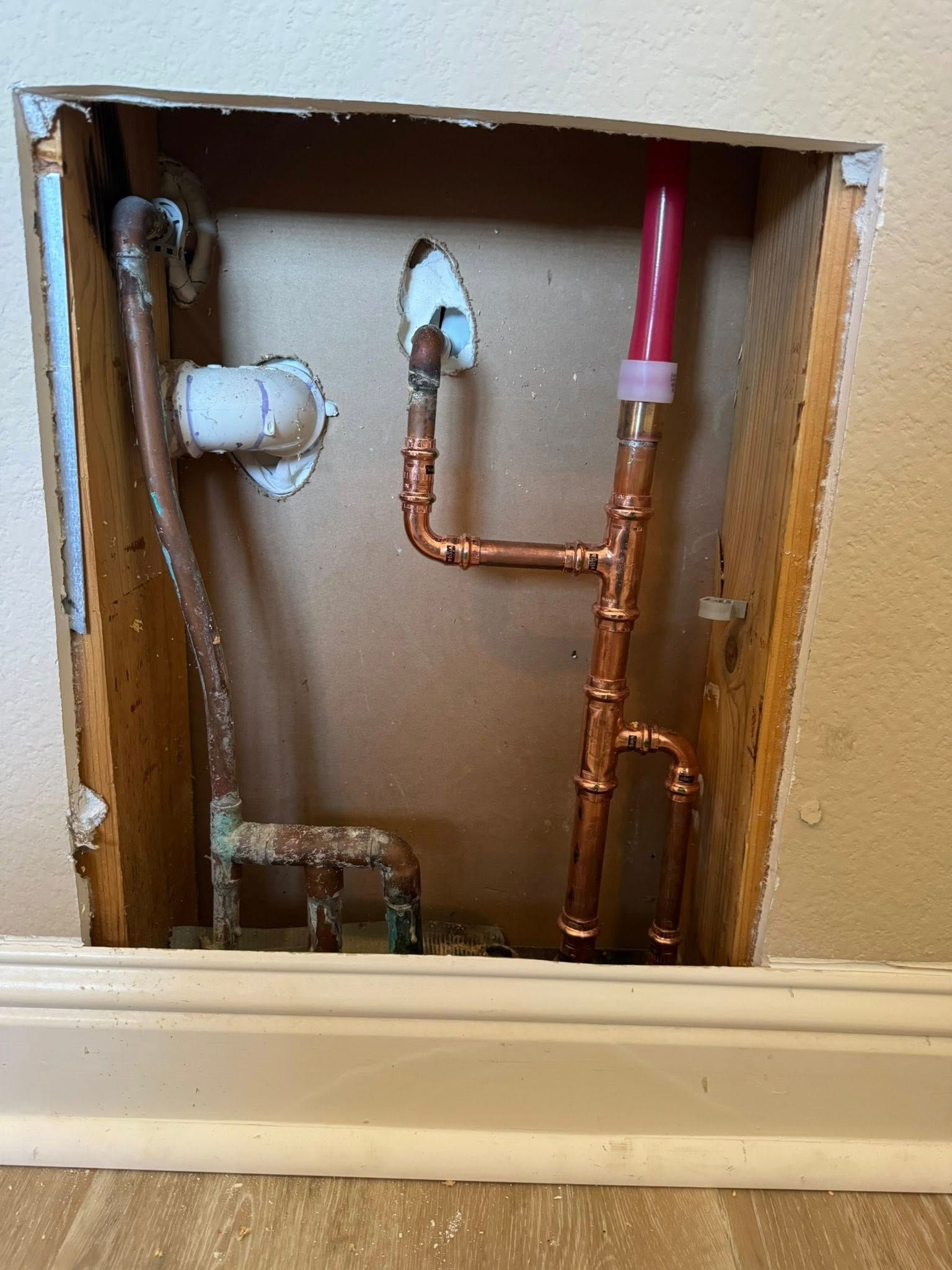 Copper pipes with a red PEX connector installed inside a hole cut into a residential wall.