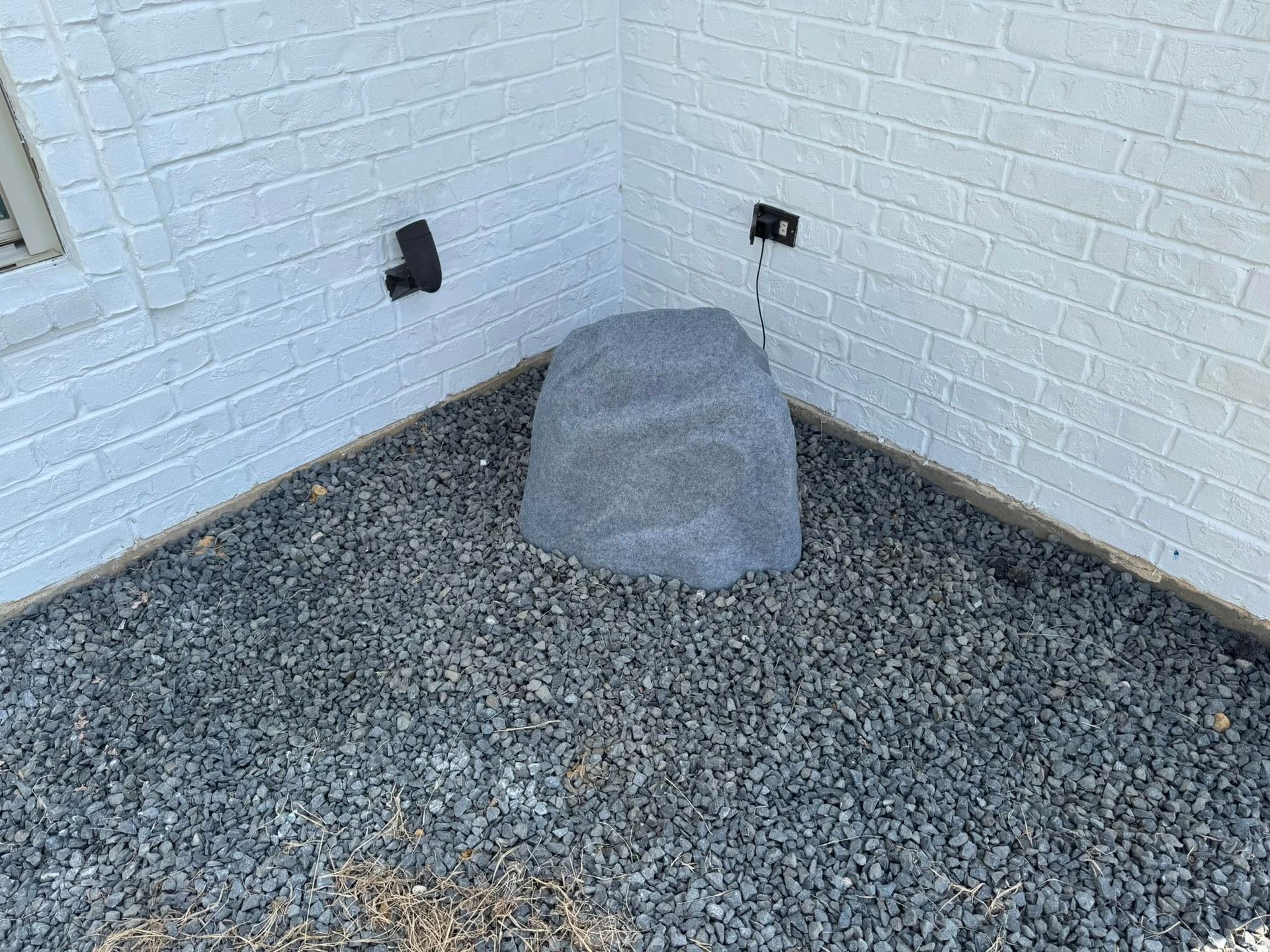 A large, grey faux-rock cover sits on a gravel patch in the corner of a white brick wall, concealing an outdoor fixture.