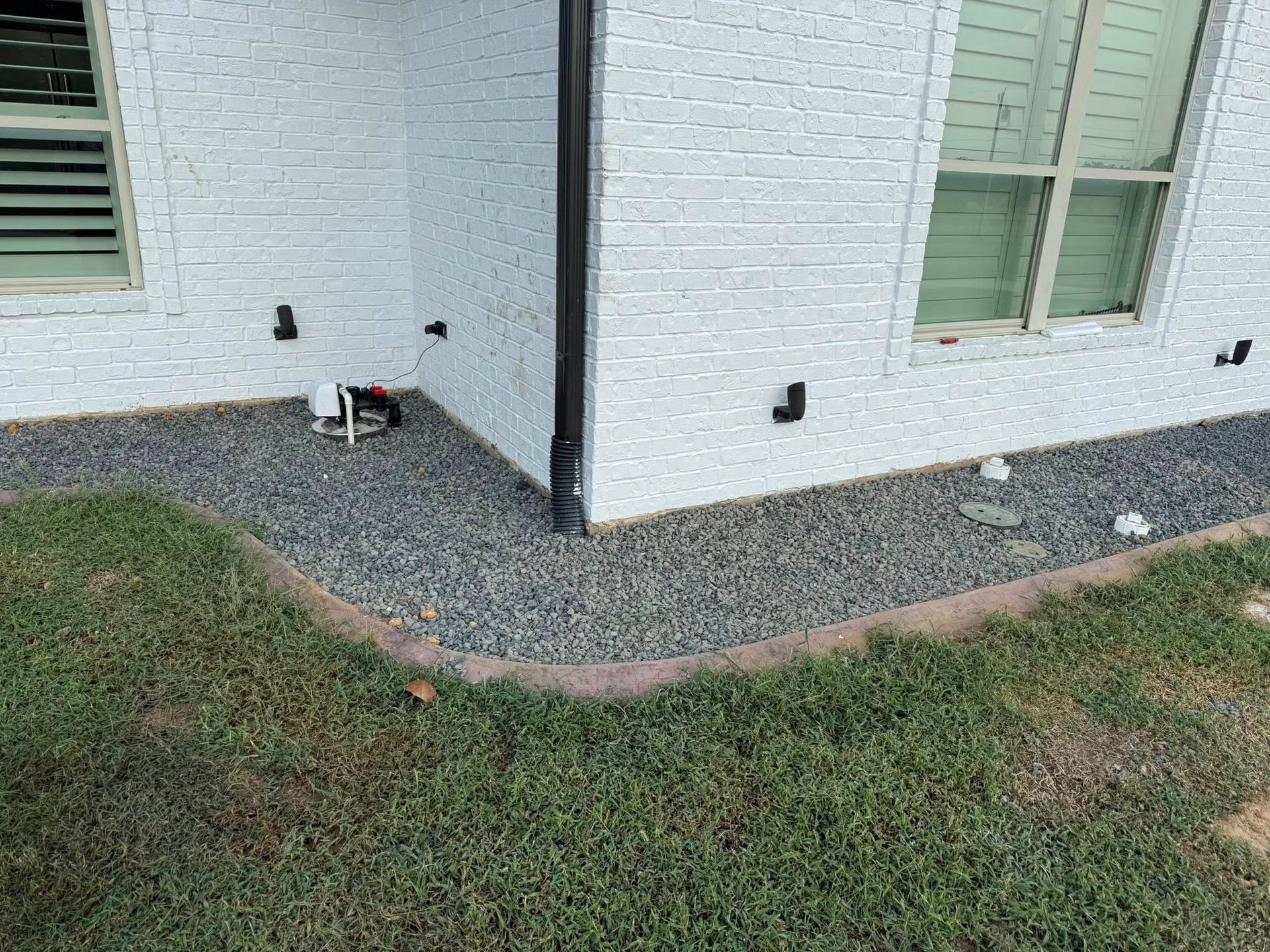 A house corner with white brick siding, a black downspout, and a gray gravel border separating the wall from the lawn.