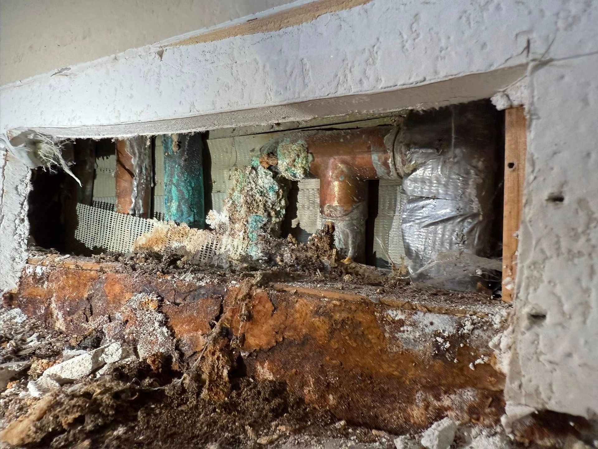 A view inside a wall showing corroded copper pipes, water-damaged wooden framing, and debris from a plumbing leak.