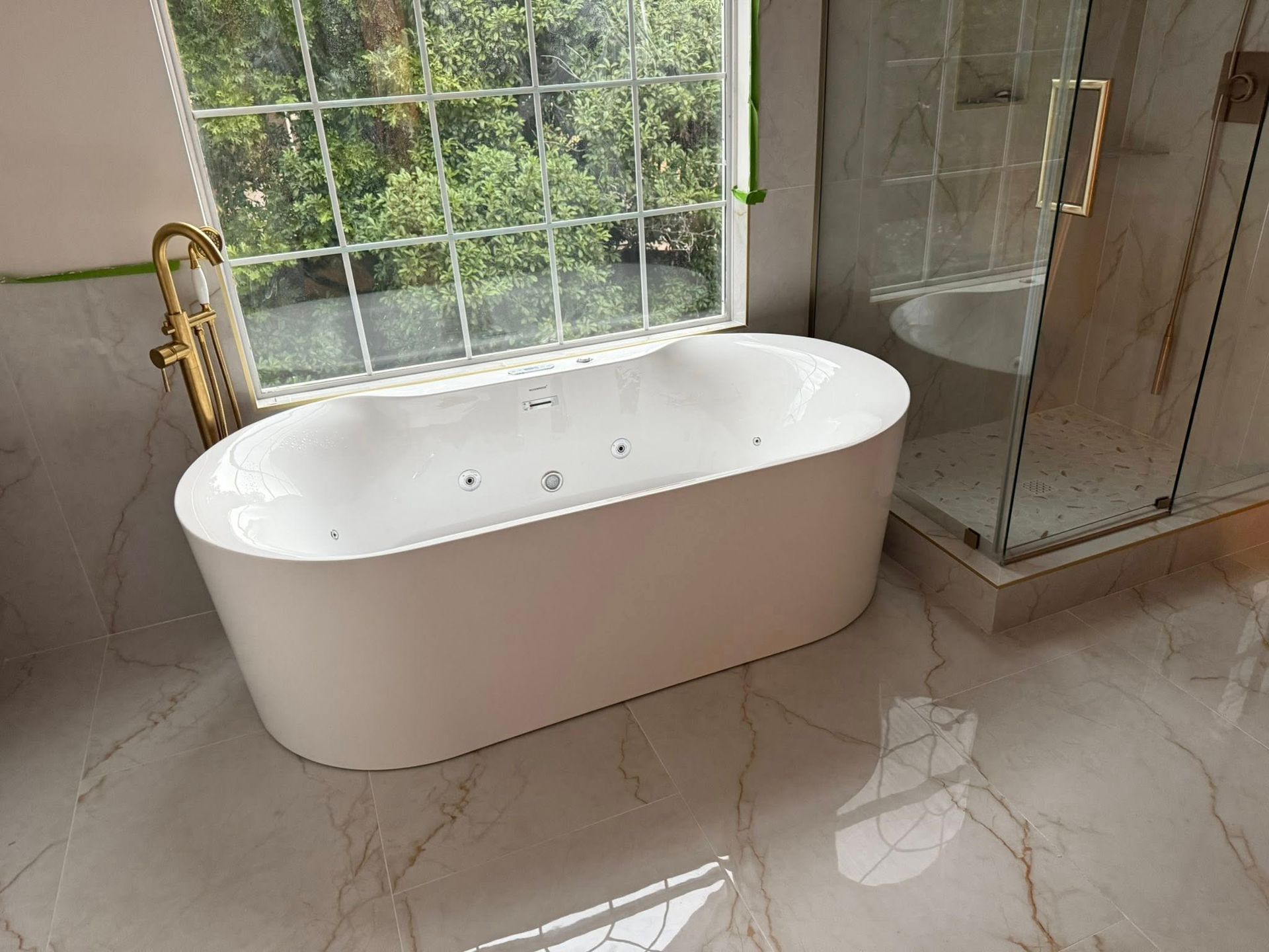 White freestanding oval jetted bathtub with a gold floor-mounted faucet in a modern bathroom with marble-look flooring.