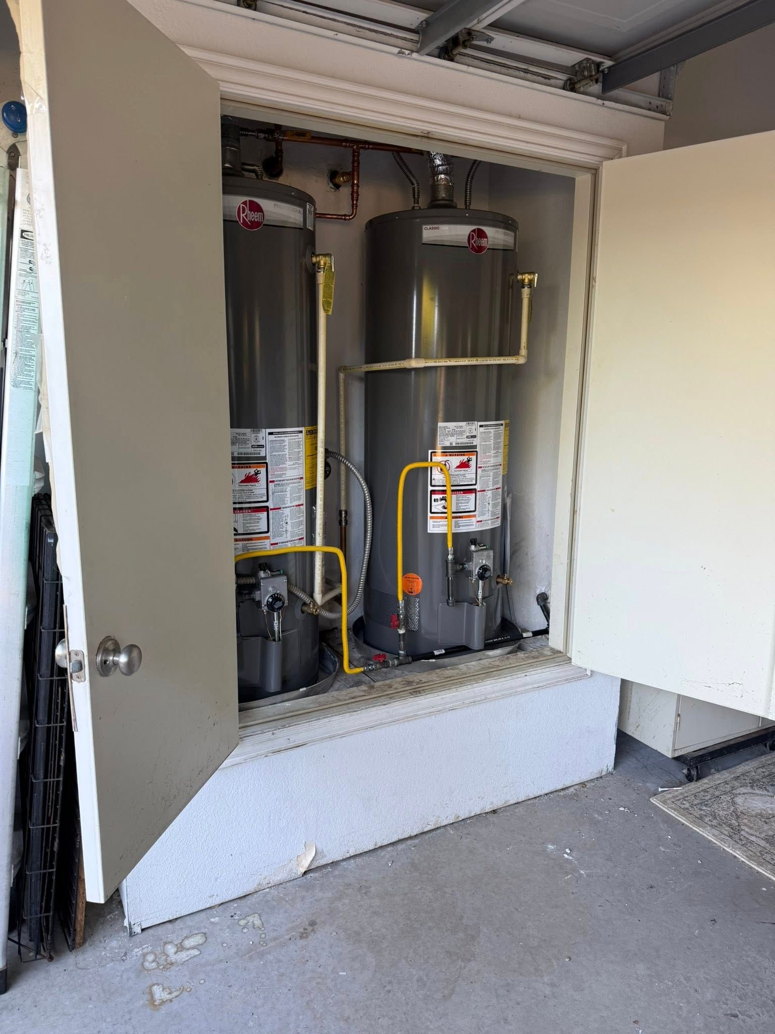 Two grey gas water heaters installed side-by-side inside an elevated garage utility closet with open doors.
