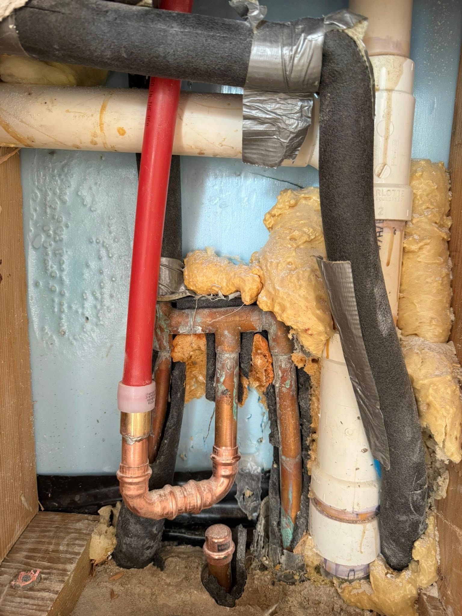 Plumbing pipes made of copper, red PEX, and white PVC, surrounded by insulation foam in a wall cavity.