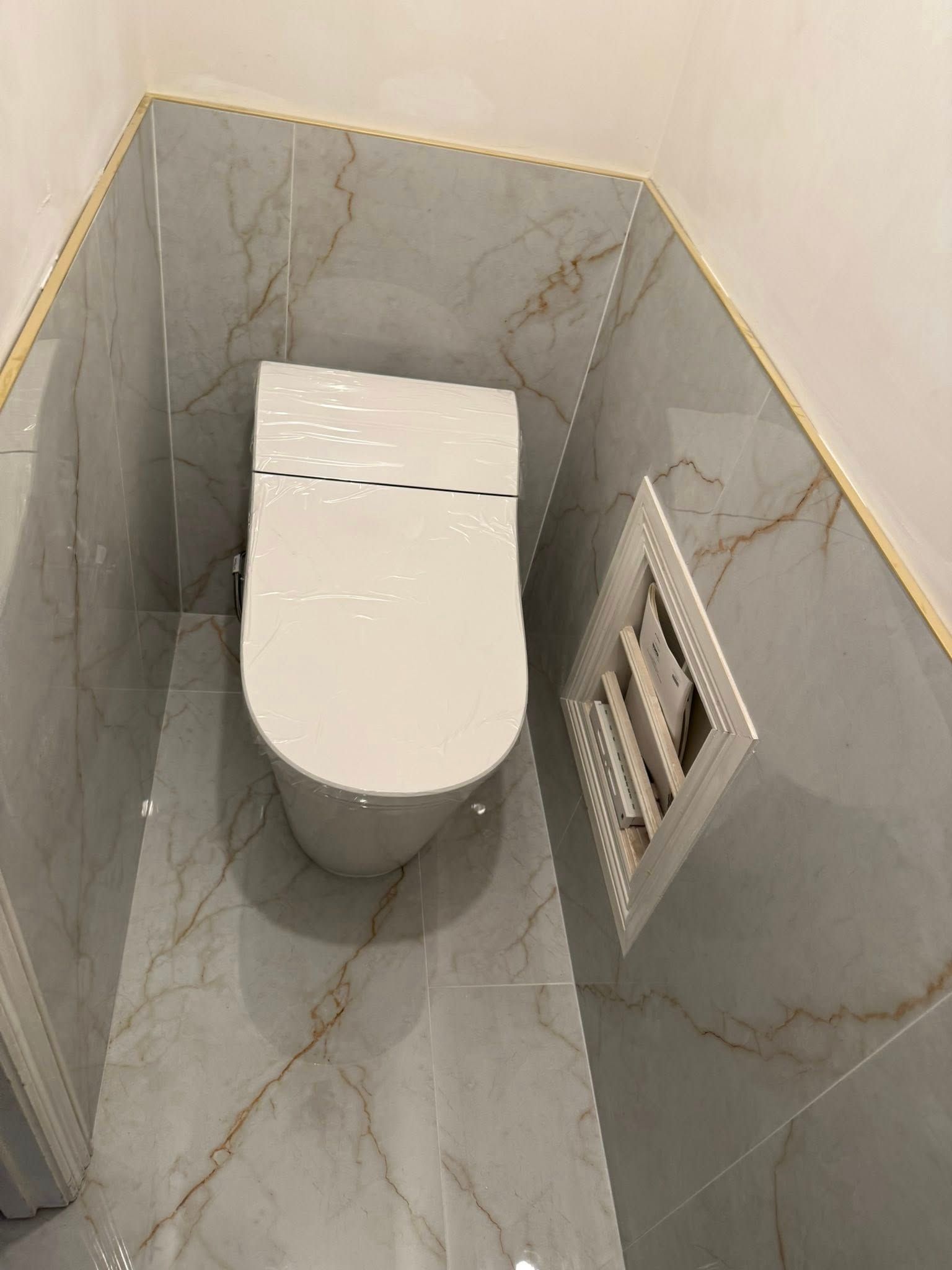A toilet in a small, tiled corner alcove with light gray, marble-patterned tiles and a white wall-access panel.