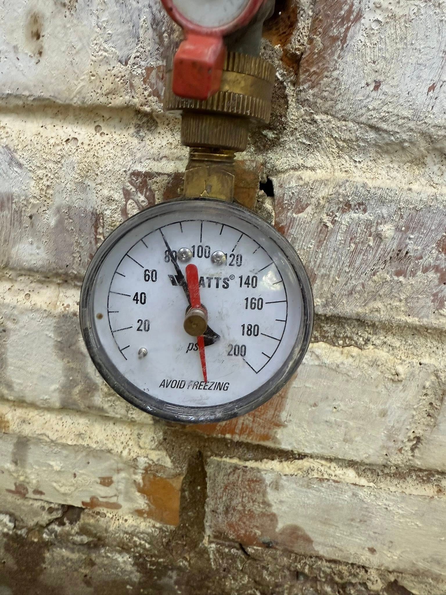 A circular pressure gauge mounted on a brick wall, with a needle pointing to approximately 80 PSI.