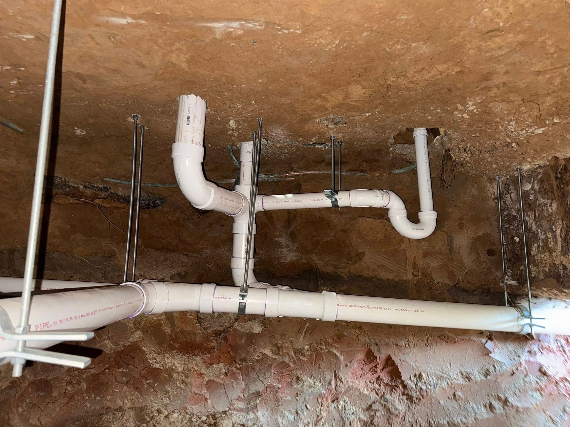 White PVC drain pipes attached to a ceiling with metal straps, running horizontally and vertically in a basement space.