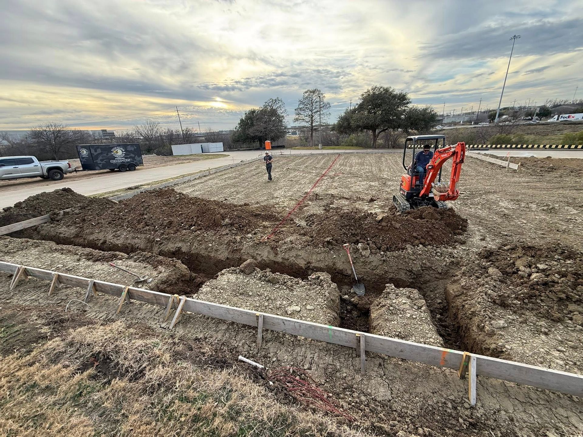An orange excavator digs a trench in an open construction site with wooden formwork visible in the foreground.