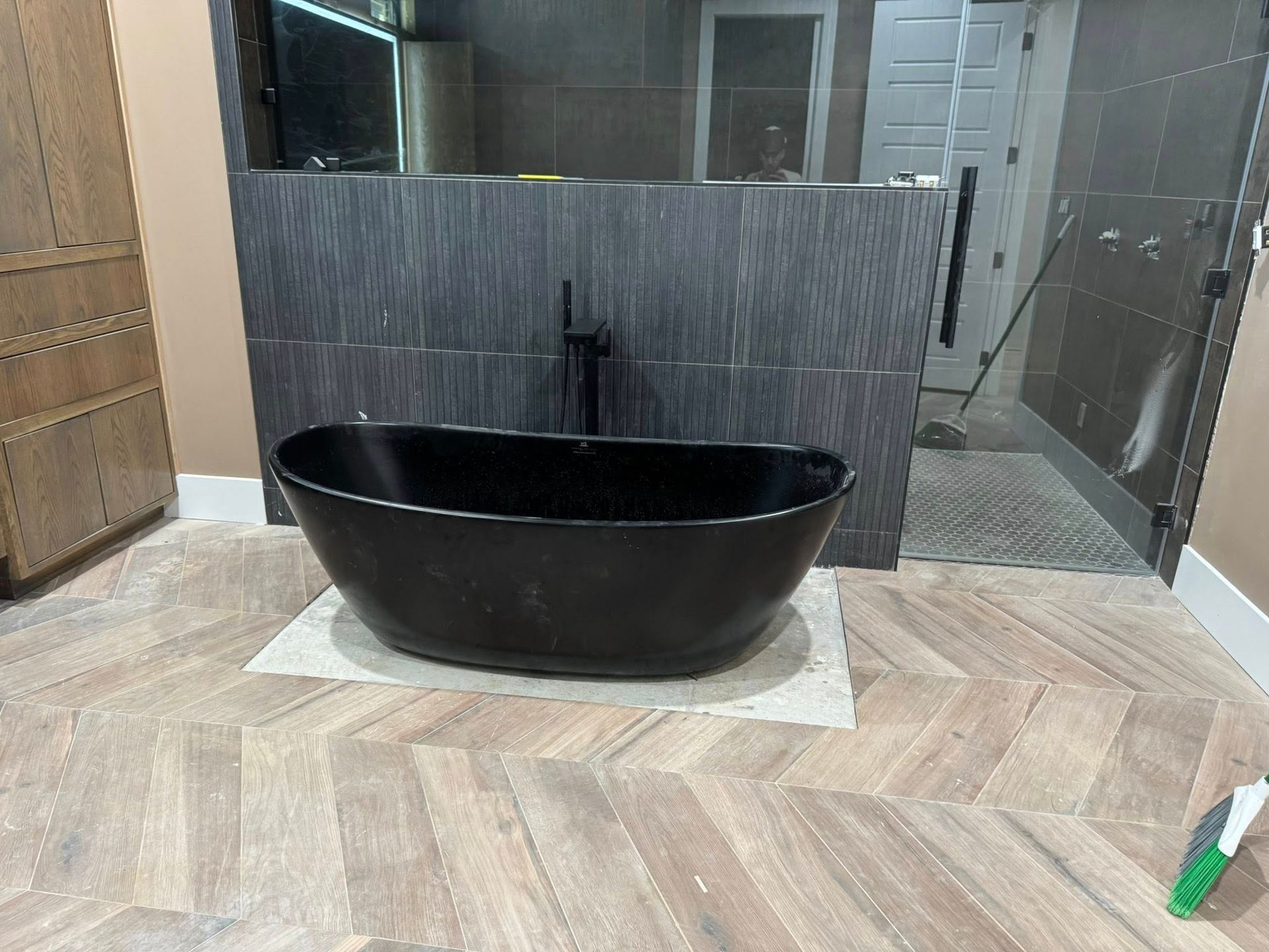 A black freestanding oval bathtub sits on a light-colored rug against a dark, textured tile wall in a modern bathroom.