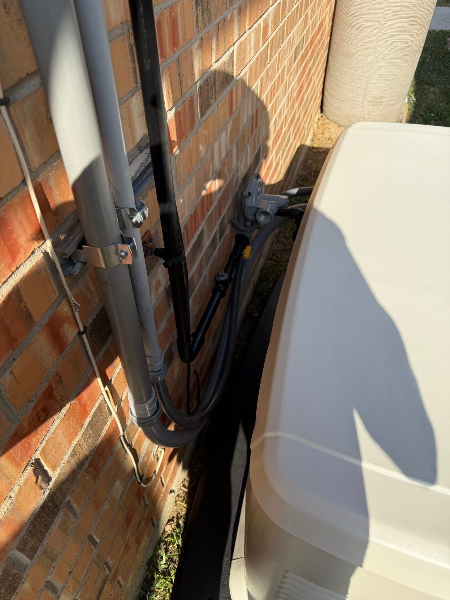 Exterior view of a residential brick wall with utility pipes, a gas shut-off valve, and part of a white generator unit.