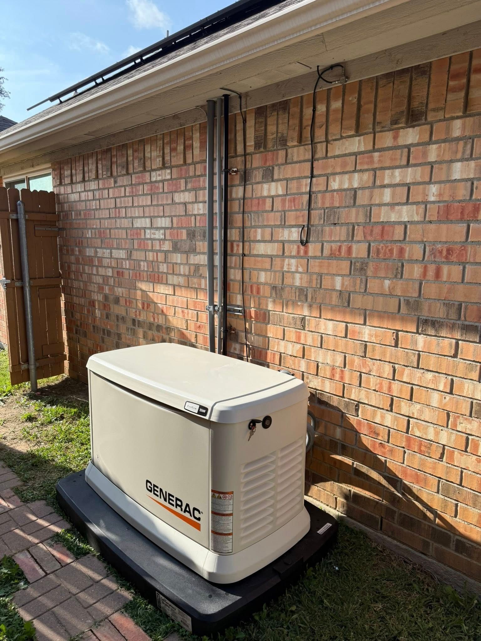 A tan Generac standby generator sitting on a black pad next to a brick wall of a house.