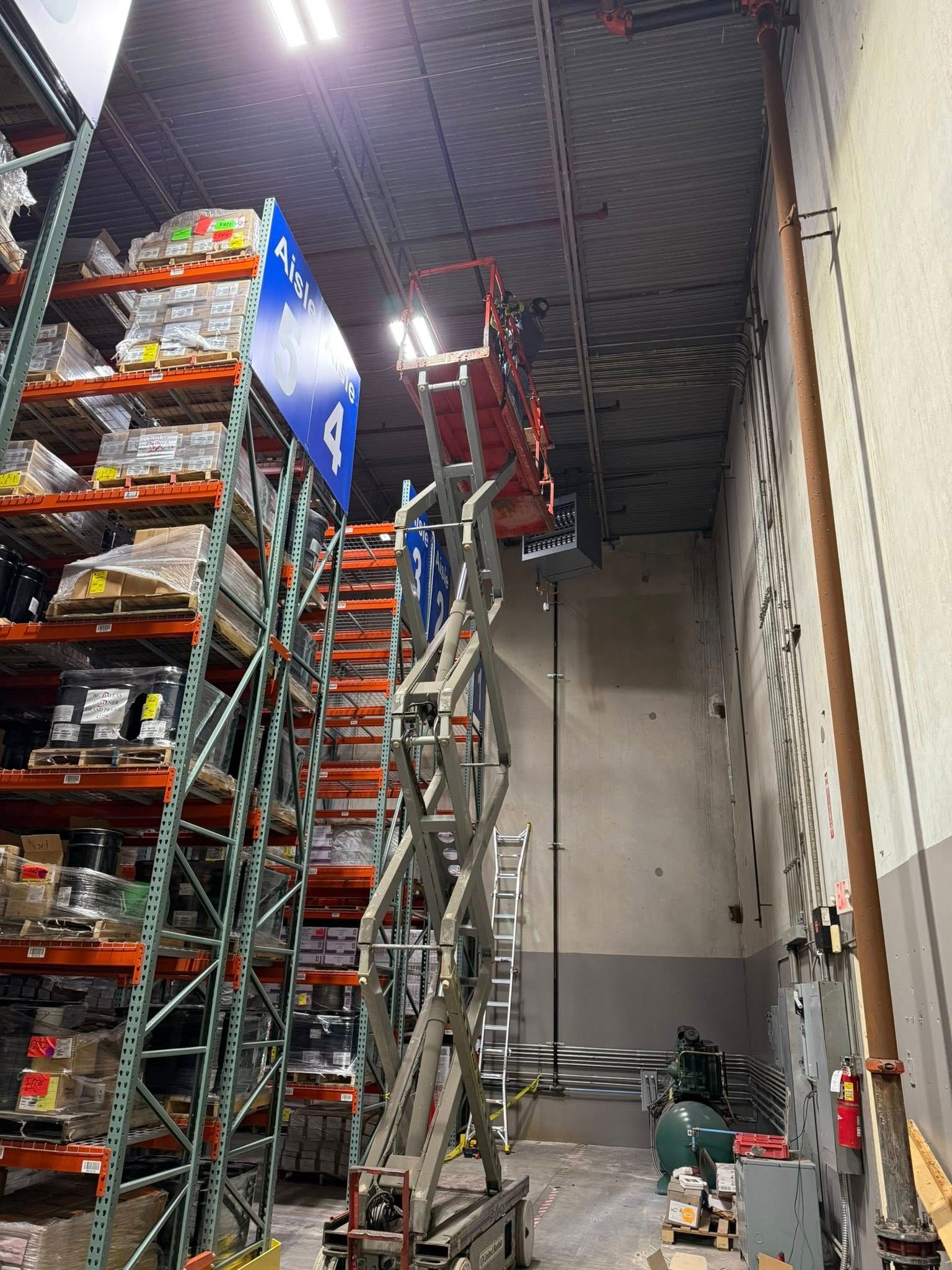 A red scissor lift extended high in a warehouse aisle next to tall metal shelving units filled with pallets of goods.