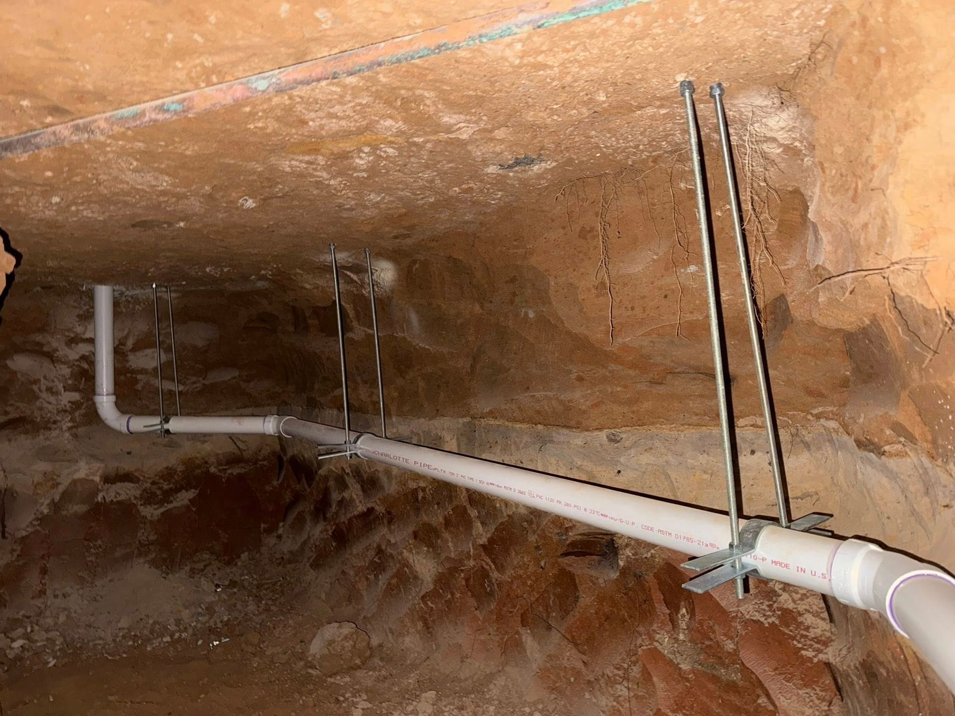 White PVC plumbing pipes suspended from a dirt ceiling by metal rods and brackets in an underground space.