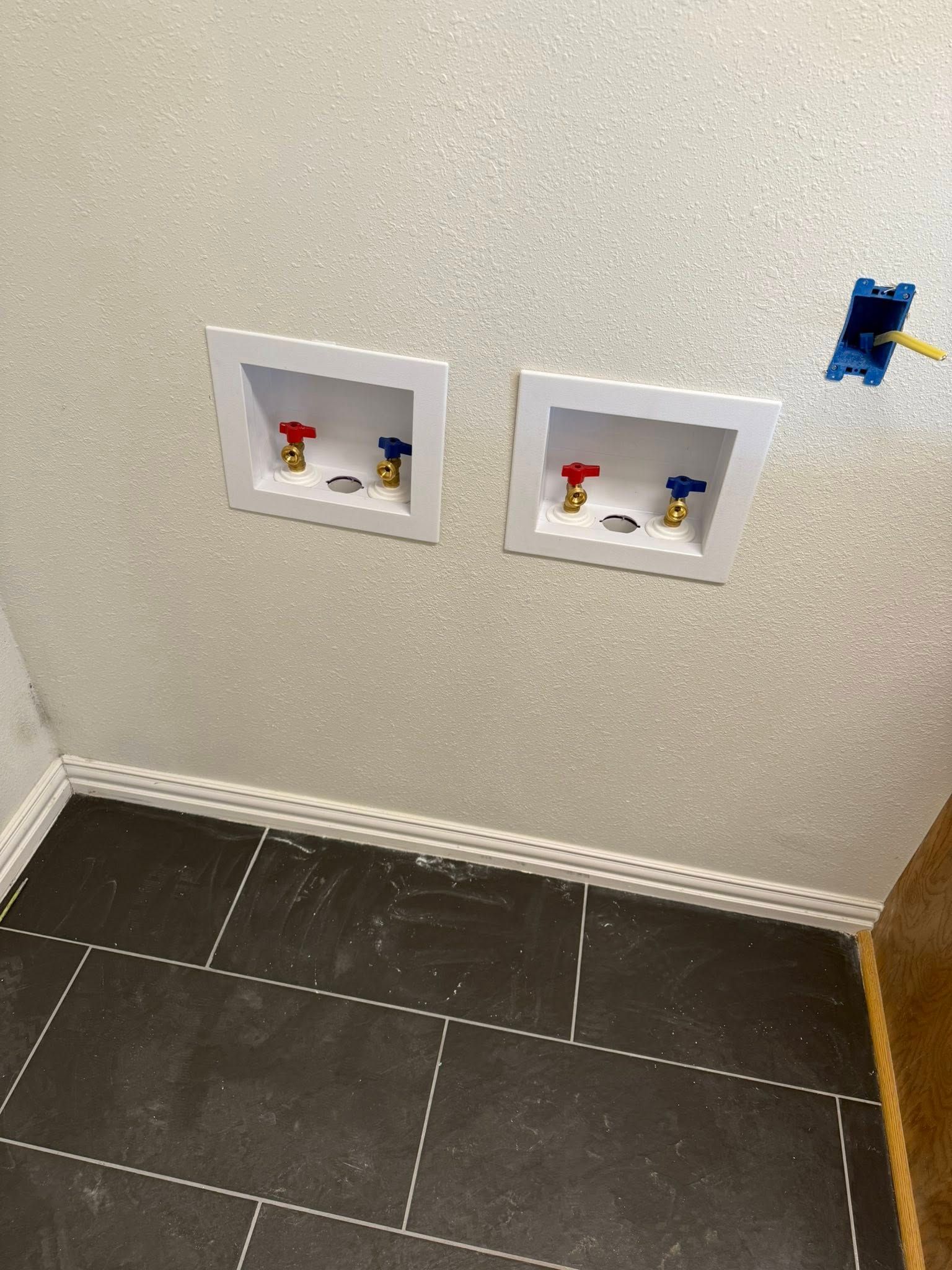 Two recessed laundry utility boxes with red and blue water valves installed on a wall above a dark tiled floor.