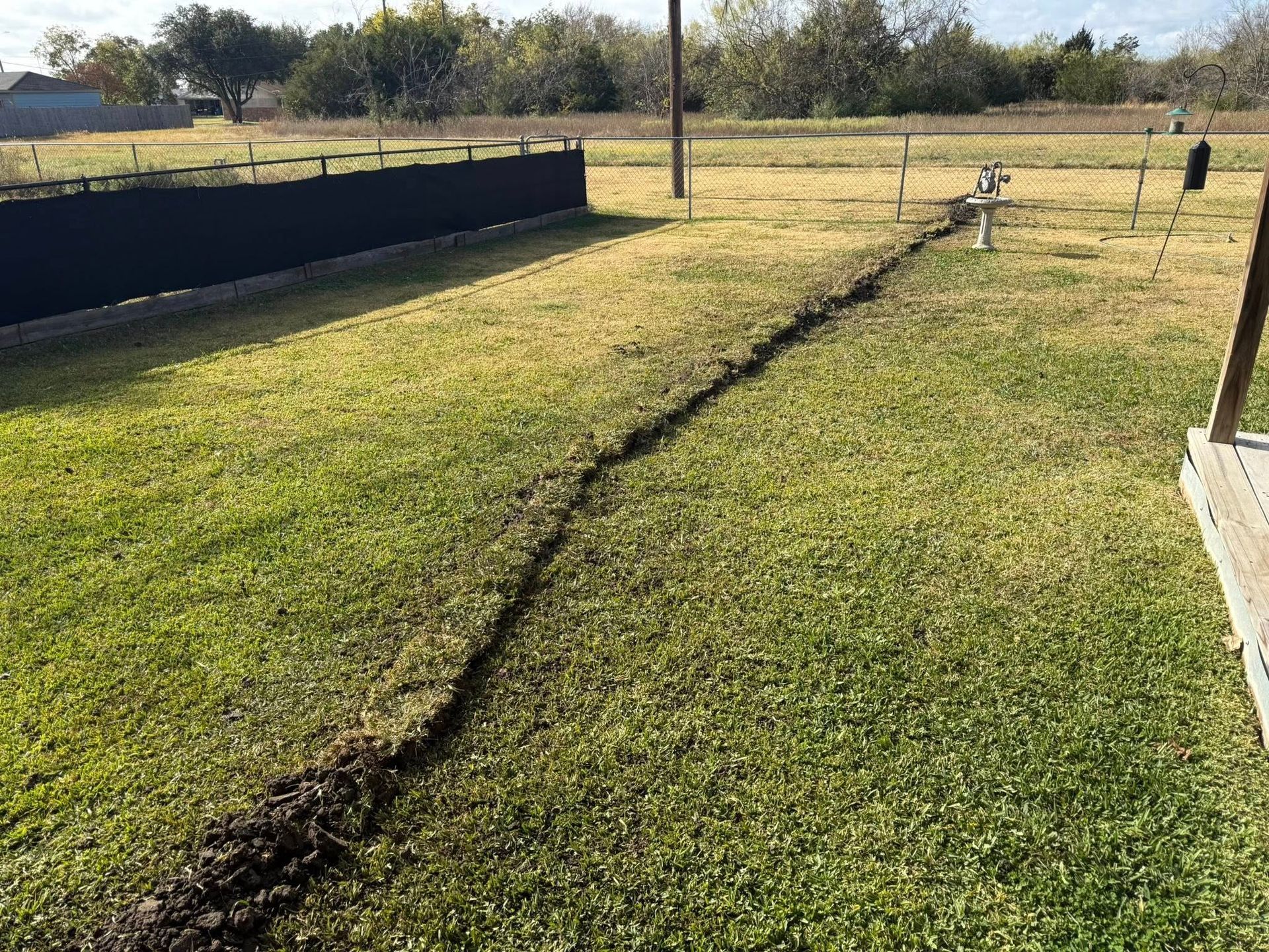 A narrow, freshly dug trench cuts through a green residential lawn, leading toward a utility pole and small faucet.