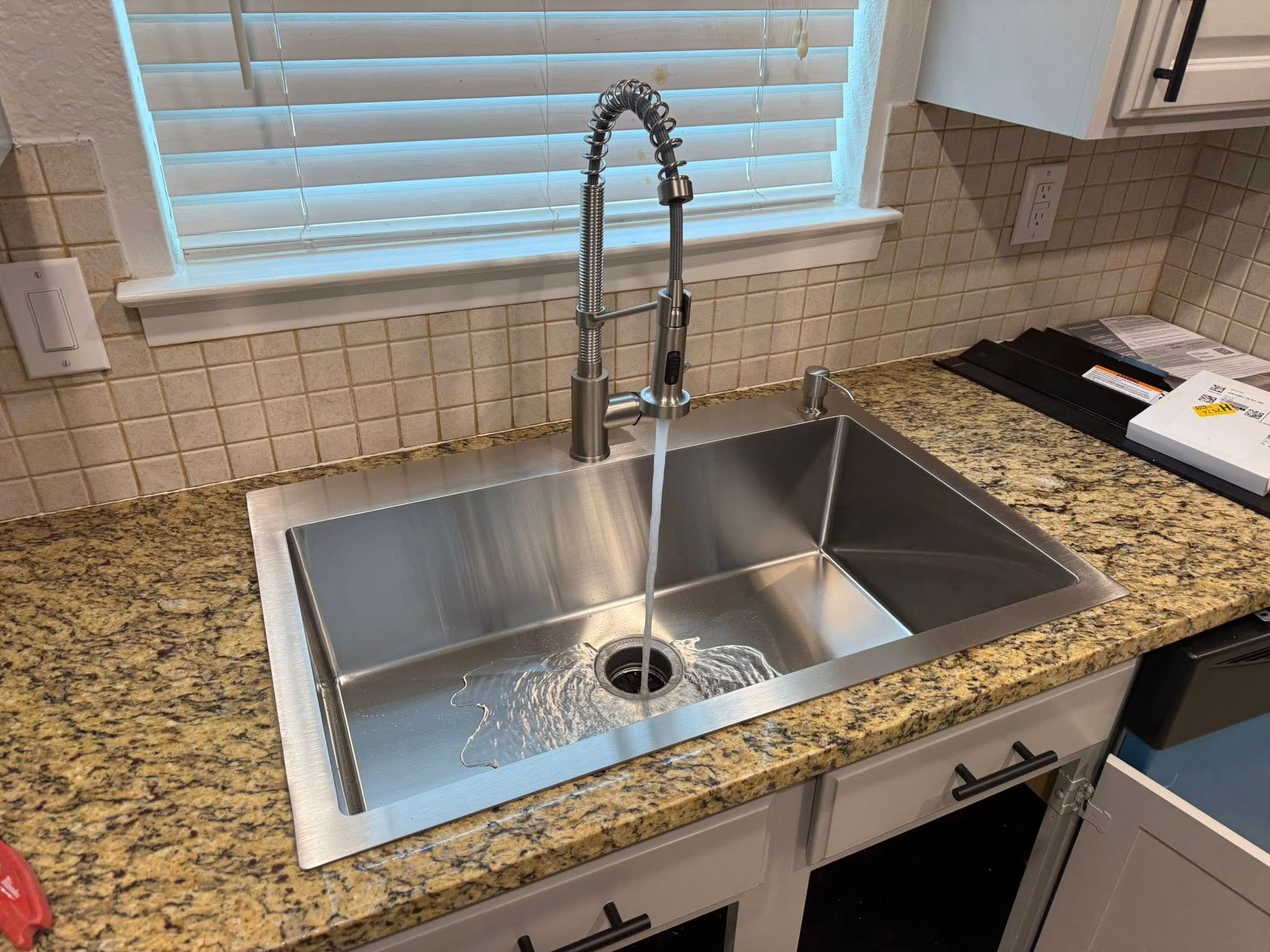 A stainless steel kitchen sink with a high-neck pull-down faucet running water into the drain, set in a granite countertop.