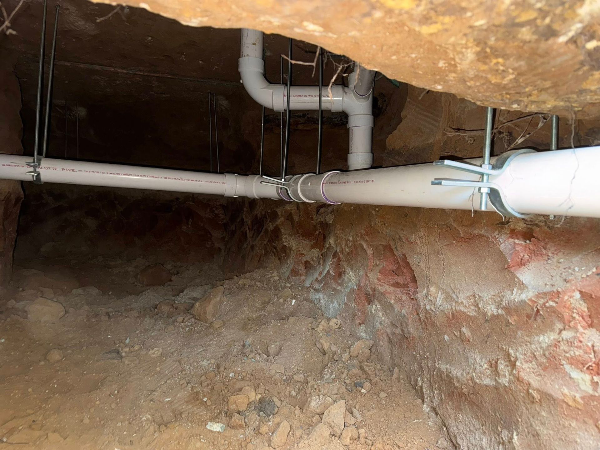 White PVC drainage pipes hang from a dirt ceiling in a crawl space, supported by metal straps.