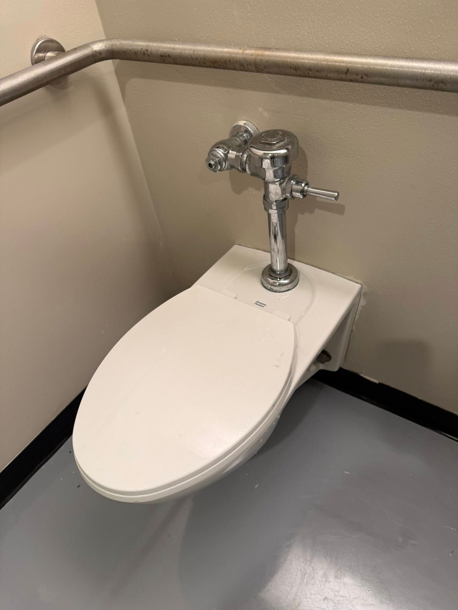 A wall-mounted toilet with a closed white seat and a chrome flush valve in a restroom with a metal grab bar.