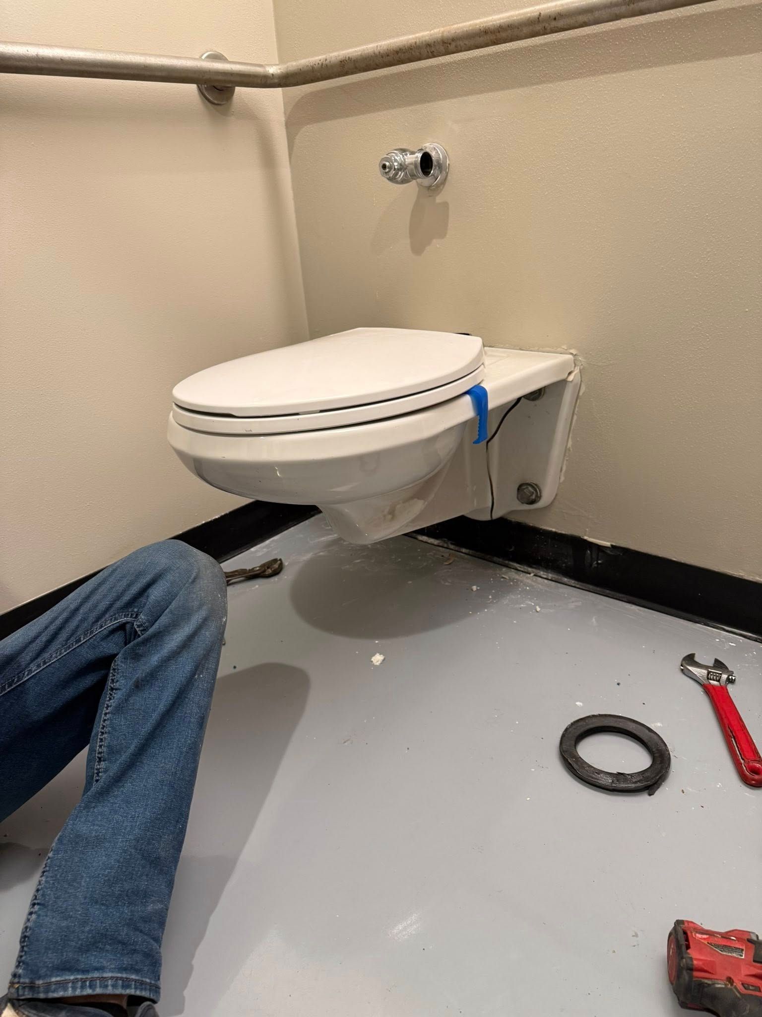 A worker is repairing a white wall-mounted toilet in a restroom. A black gasket and tools are on the floor nearby.