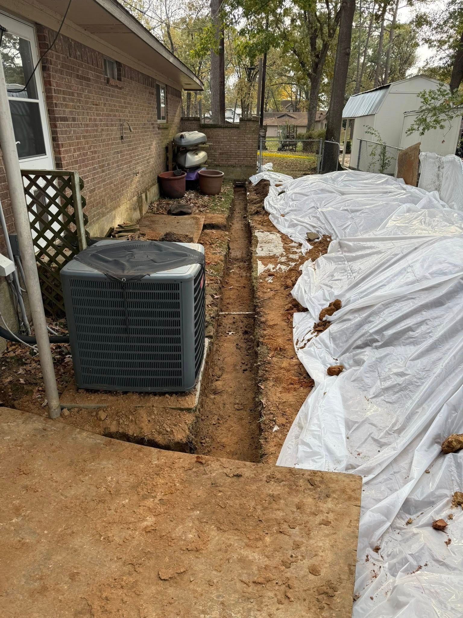 A narrow trench dug in the dirt alongside a brick house exterior, with an AC unit and a white plastic sheet nearby.