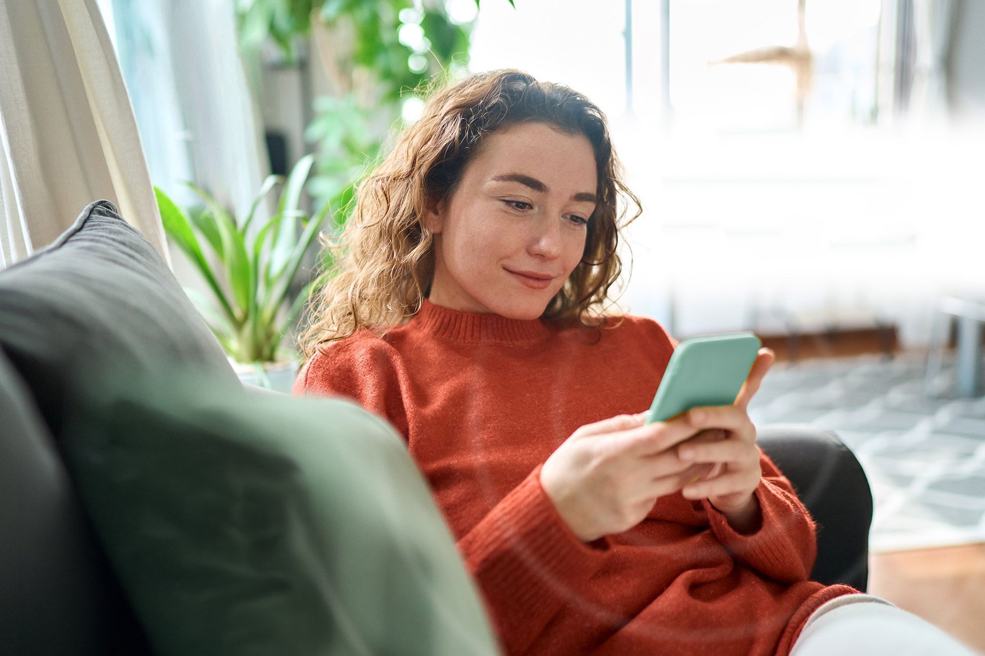 Woman in an orange sweater sitting on a couch using her smartphone and the oto app.