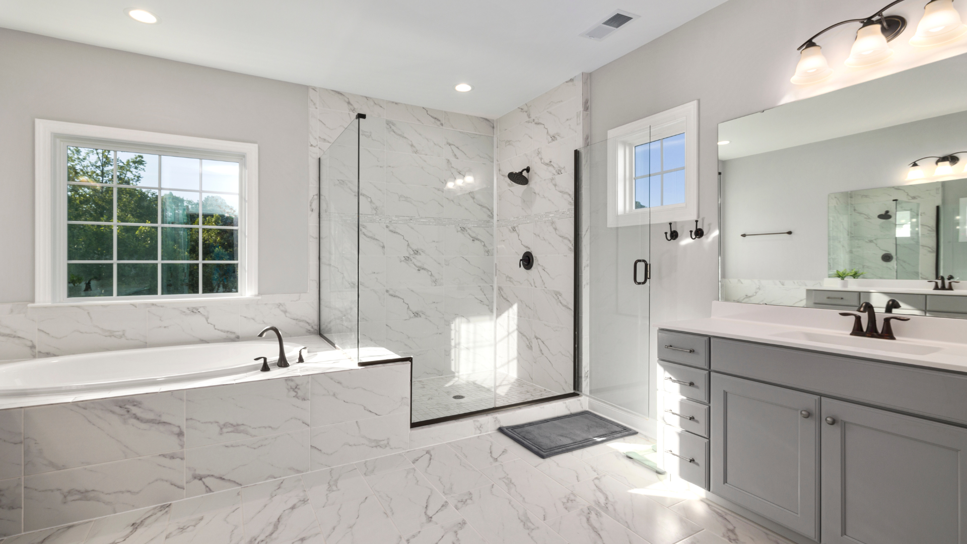 Bright, modern bathroom with marble tile, glass shower, soaking tub, vanity, and window.