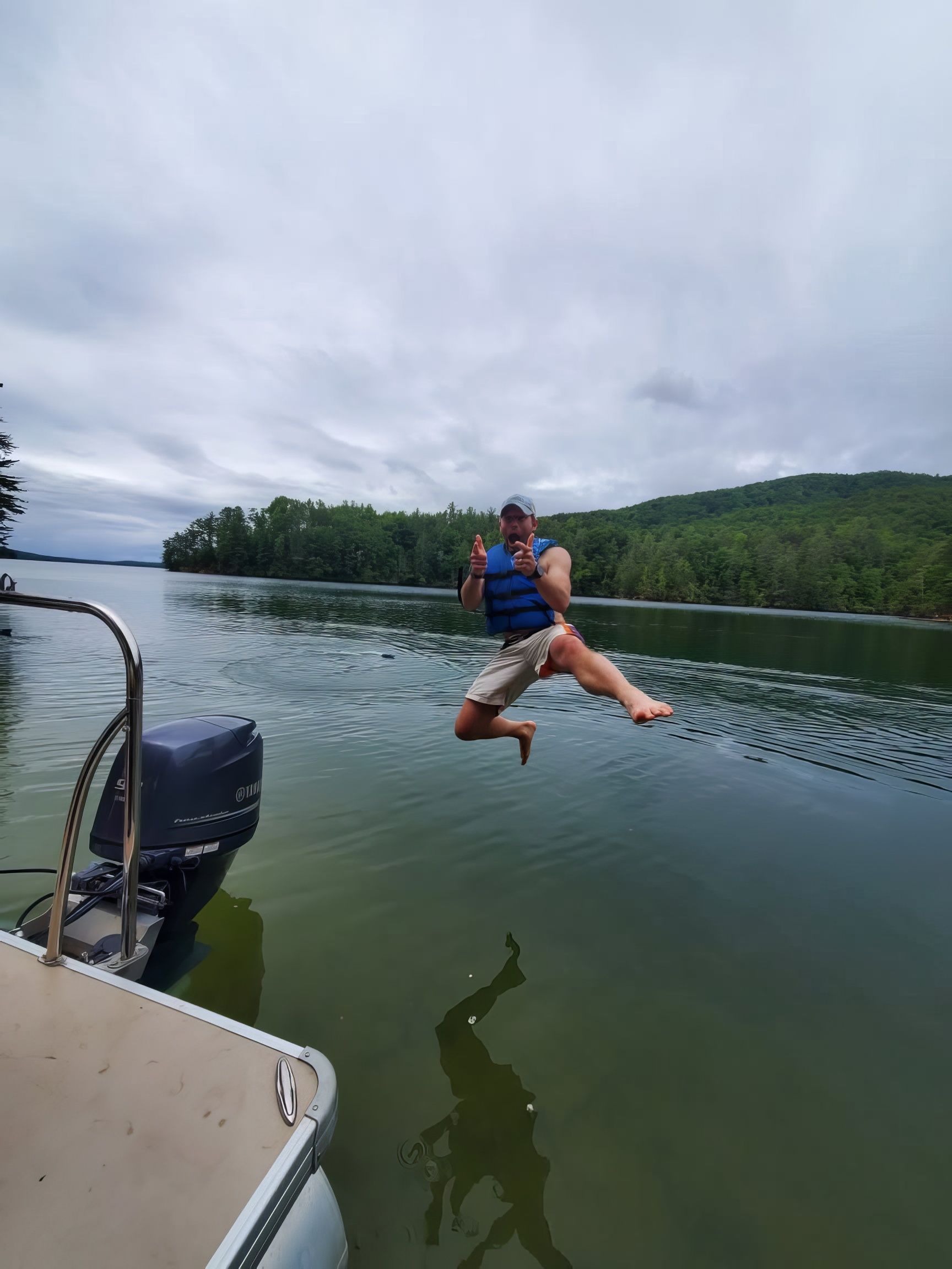 Person in a blue life jacket jumping into a lake beside a boat on an overcast day
