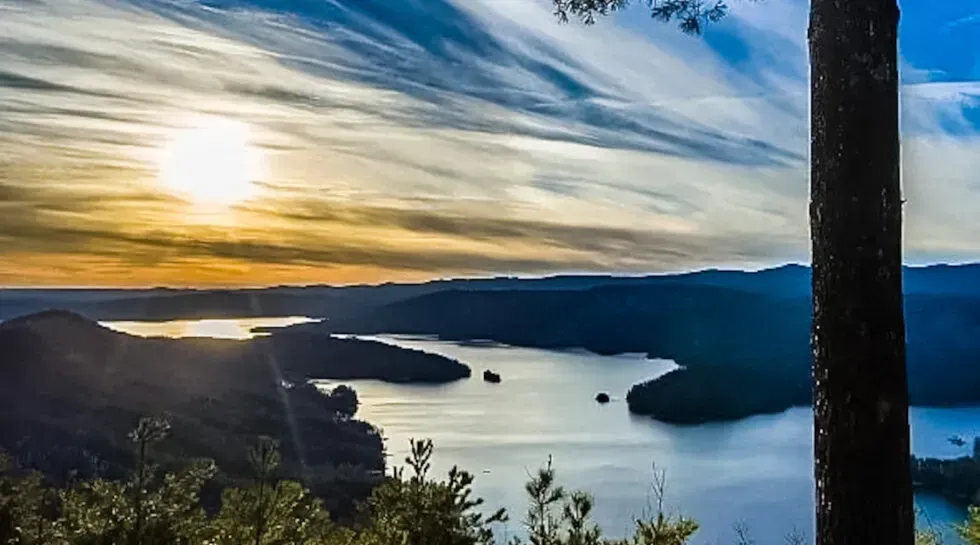 Sunset over a winding lake with dark hills and a cloudy sky, viewed from a hillside overlook