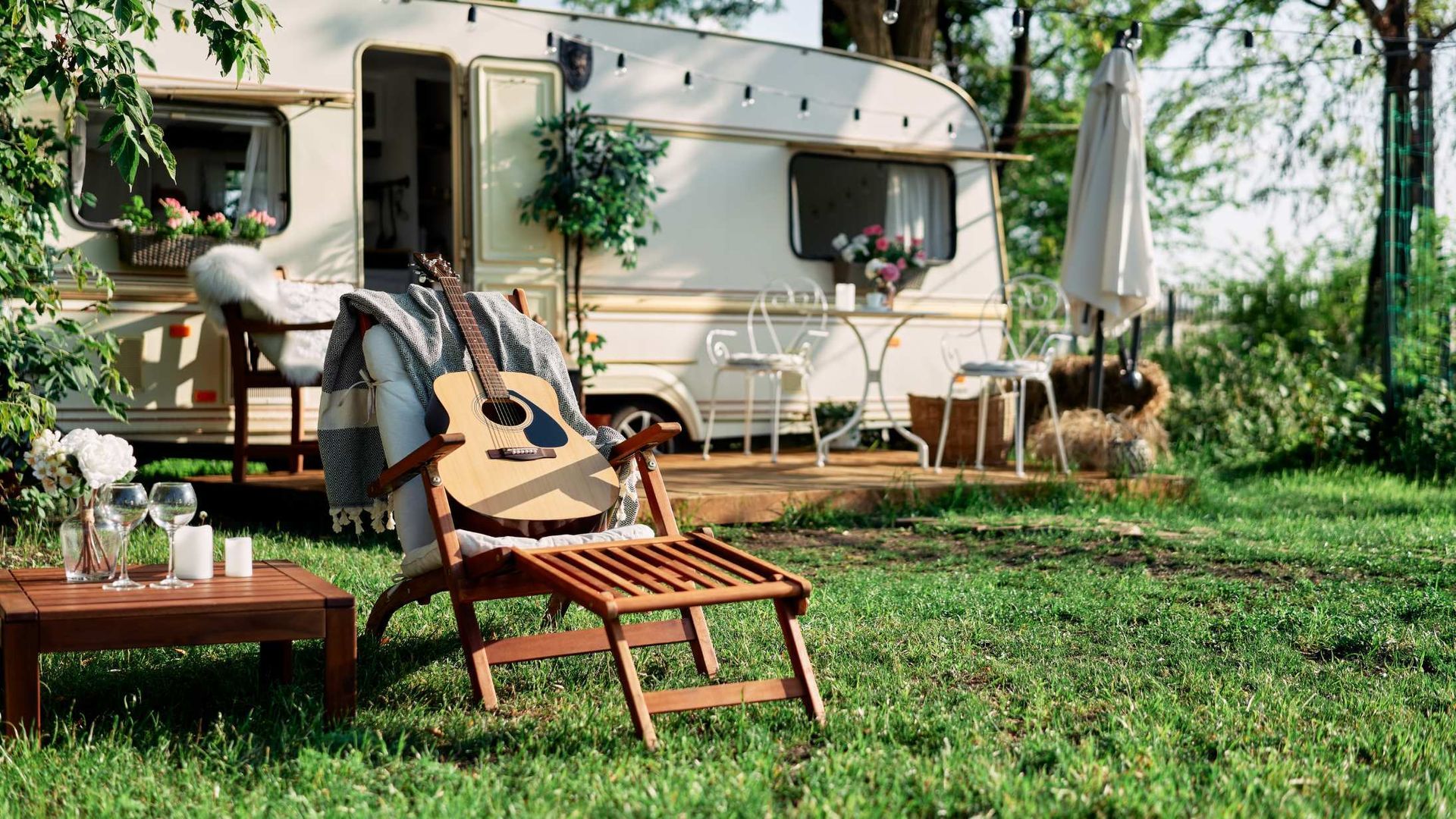 Cozy camper trailer in a grassy yard with a chair, guitar, and small patio under trees