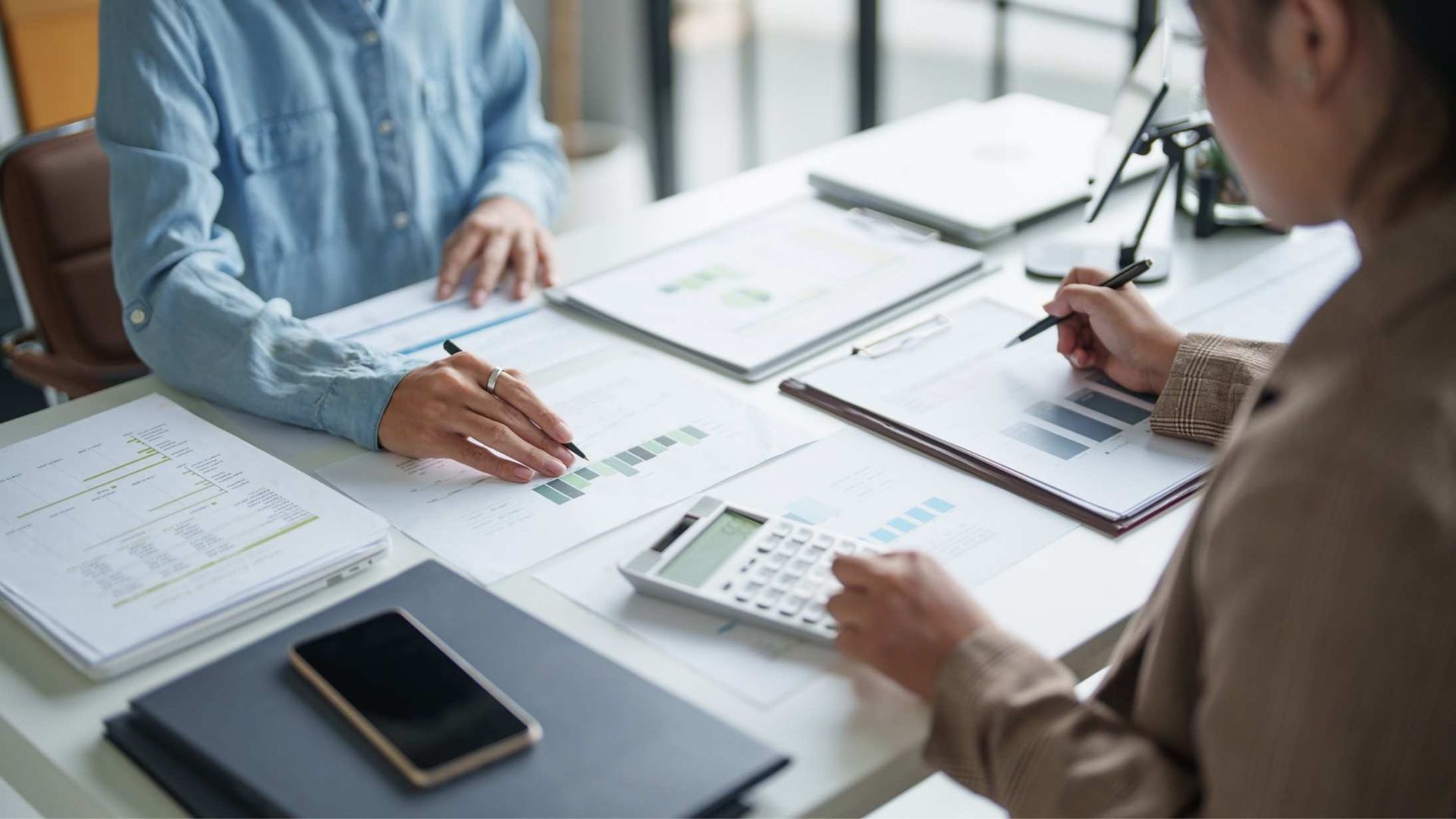 Two people reviewing documents and writing at a white office table with a smartphone and calculator