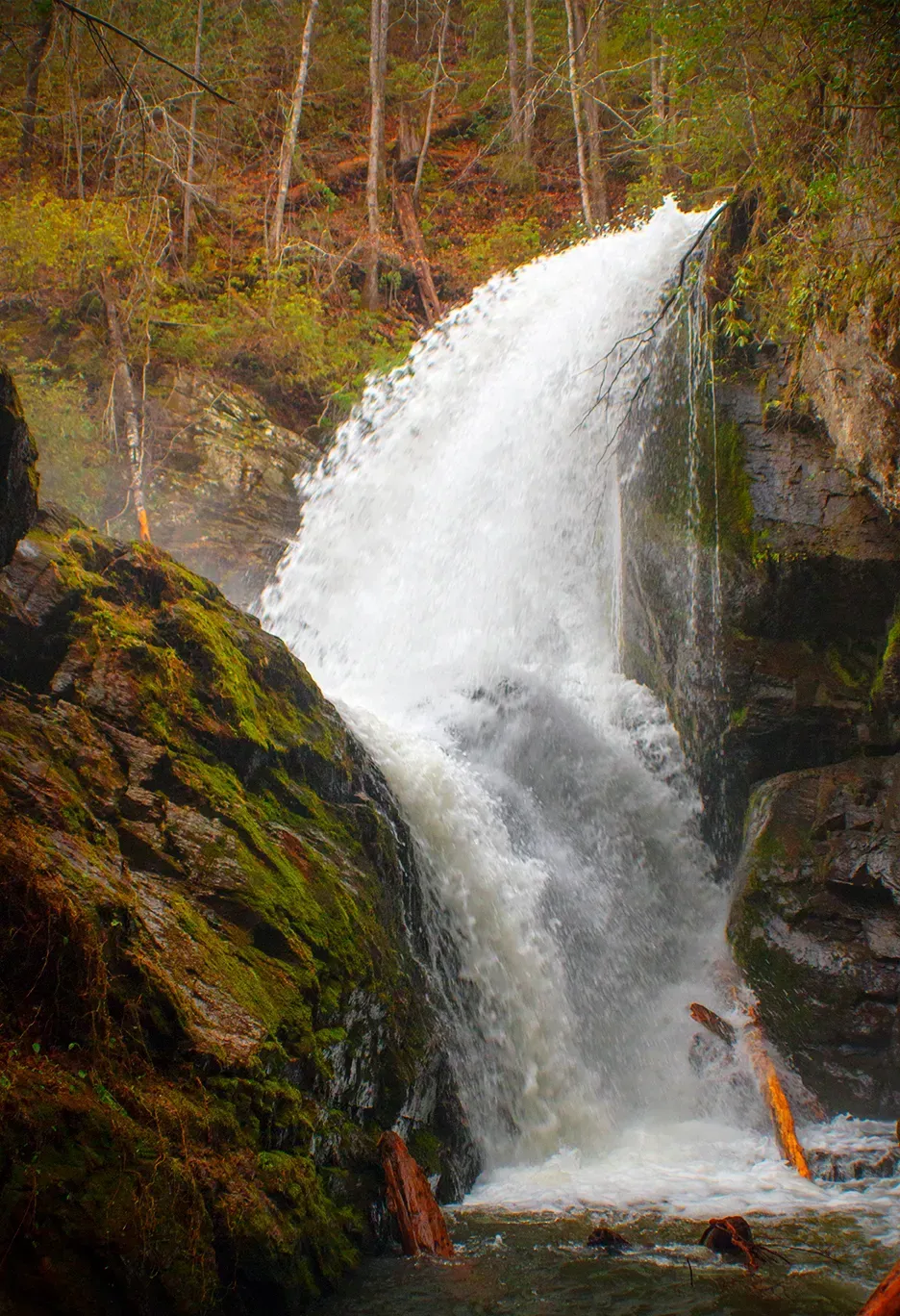 Tall white waterfall cascading over dark rocks in a forested autumn landscape